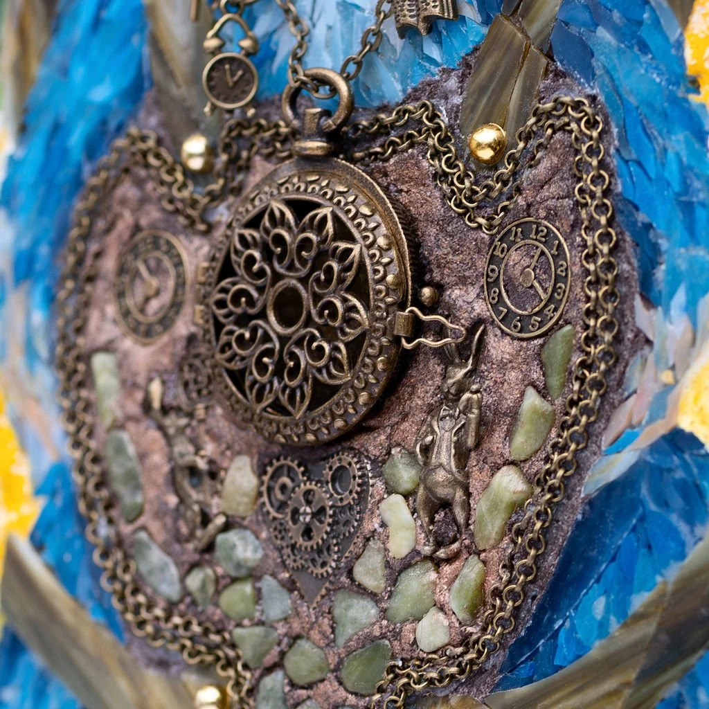 Close-up of a textured heart-shaped art piece with various chains, gears, clocks, and small statues, decorated with stones and surrounded by colorful materials.