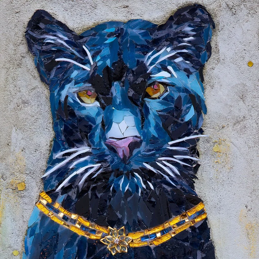 Mosaic wall art of a black and blue panther with yellow eyes, adorned with gold jewelry and a flower pendant, on a textured wall.