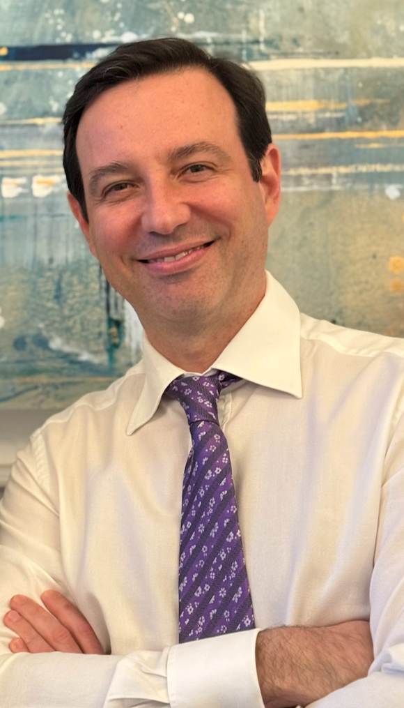 A man with dark hair, wearing a white dress shirt and a purple patterned tie, smiling with arms crossed against a blue abstract background.