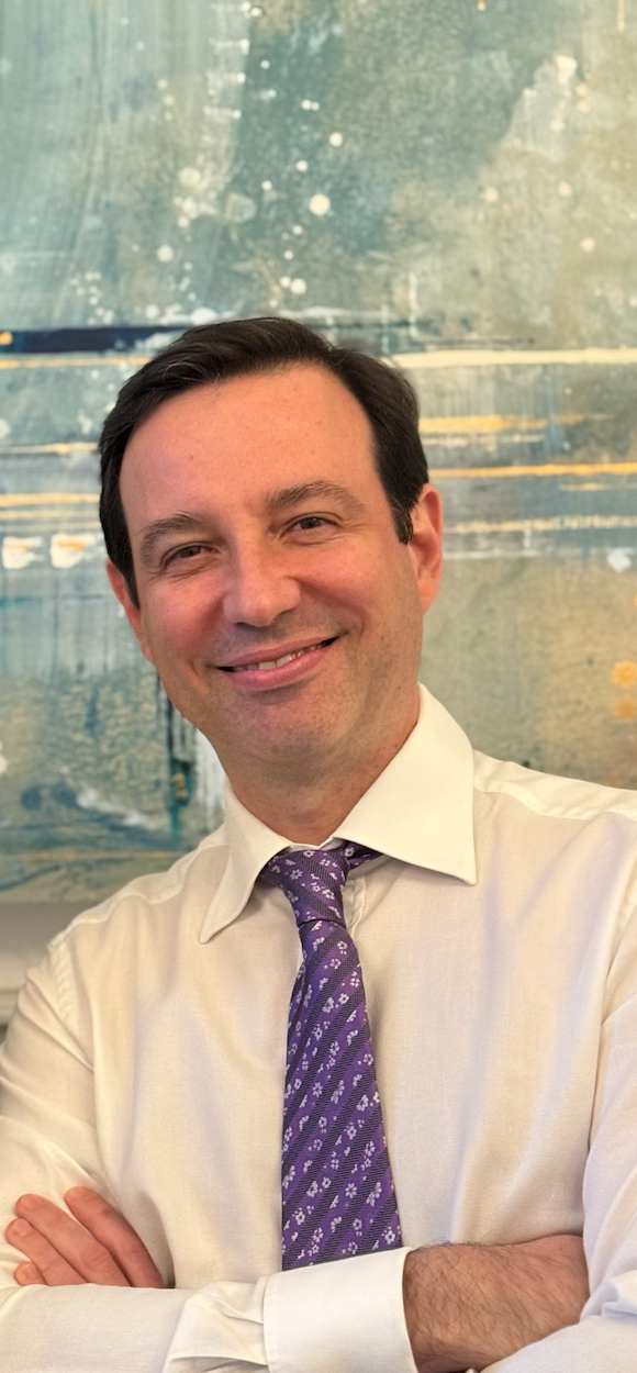 A man smiling with arms crossed, wearing a white dress shirt and a purple tie, standing in front of an abstract blue and gold background.