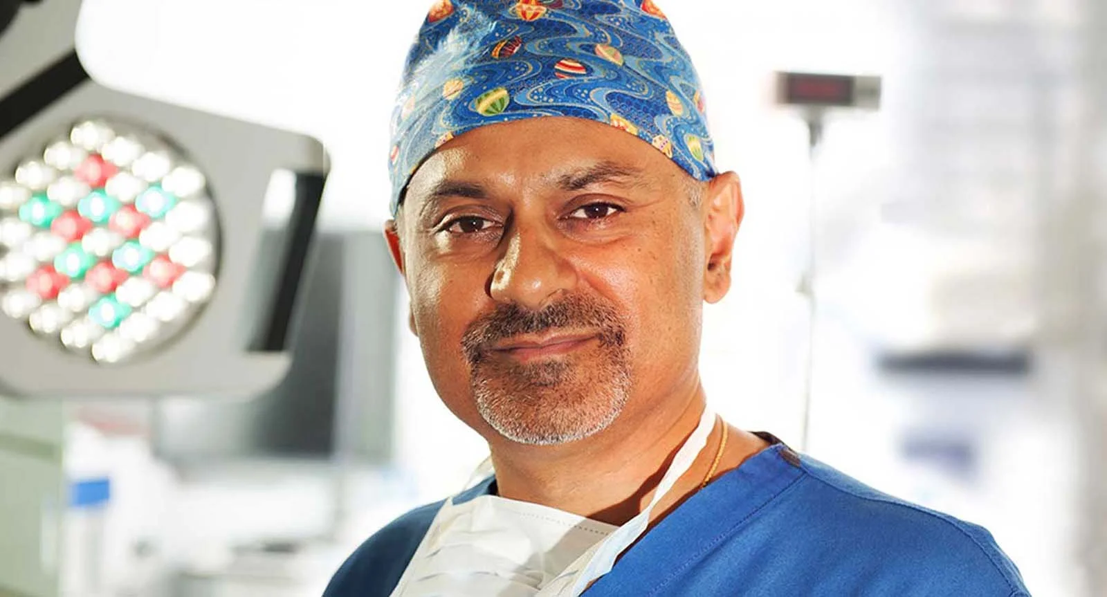 A male surgeon in blue scrubs and a patterned surgical cap standing in an operating room, with medical lighting and equipment in the background.
