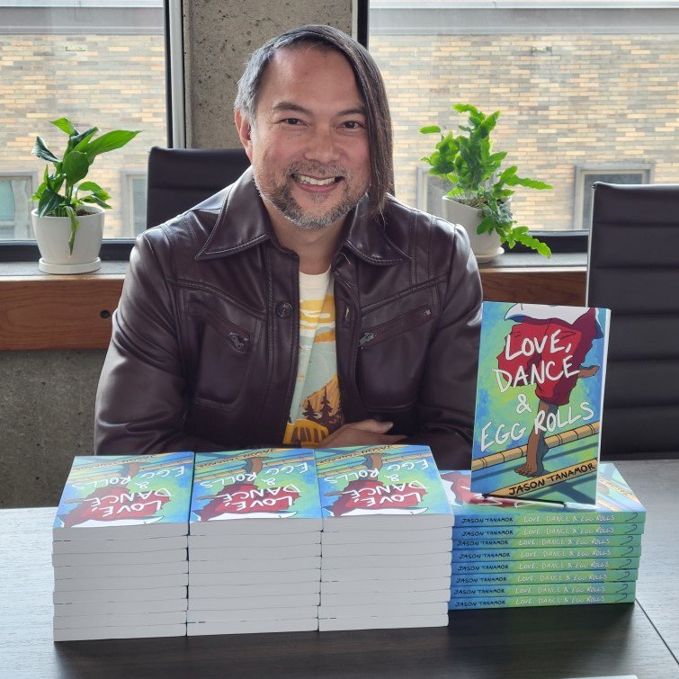 A man with long hair, a beard, and a brown leather jacket is sitting at a table with a stack of books titled "Love, Dance & Egg Rolls" by Jason Tanamor. There are two potted plants and a window in the background.