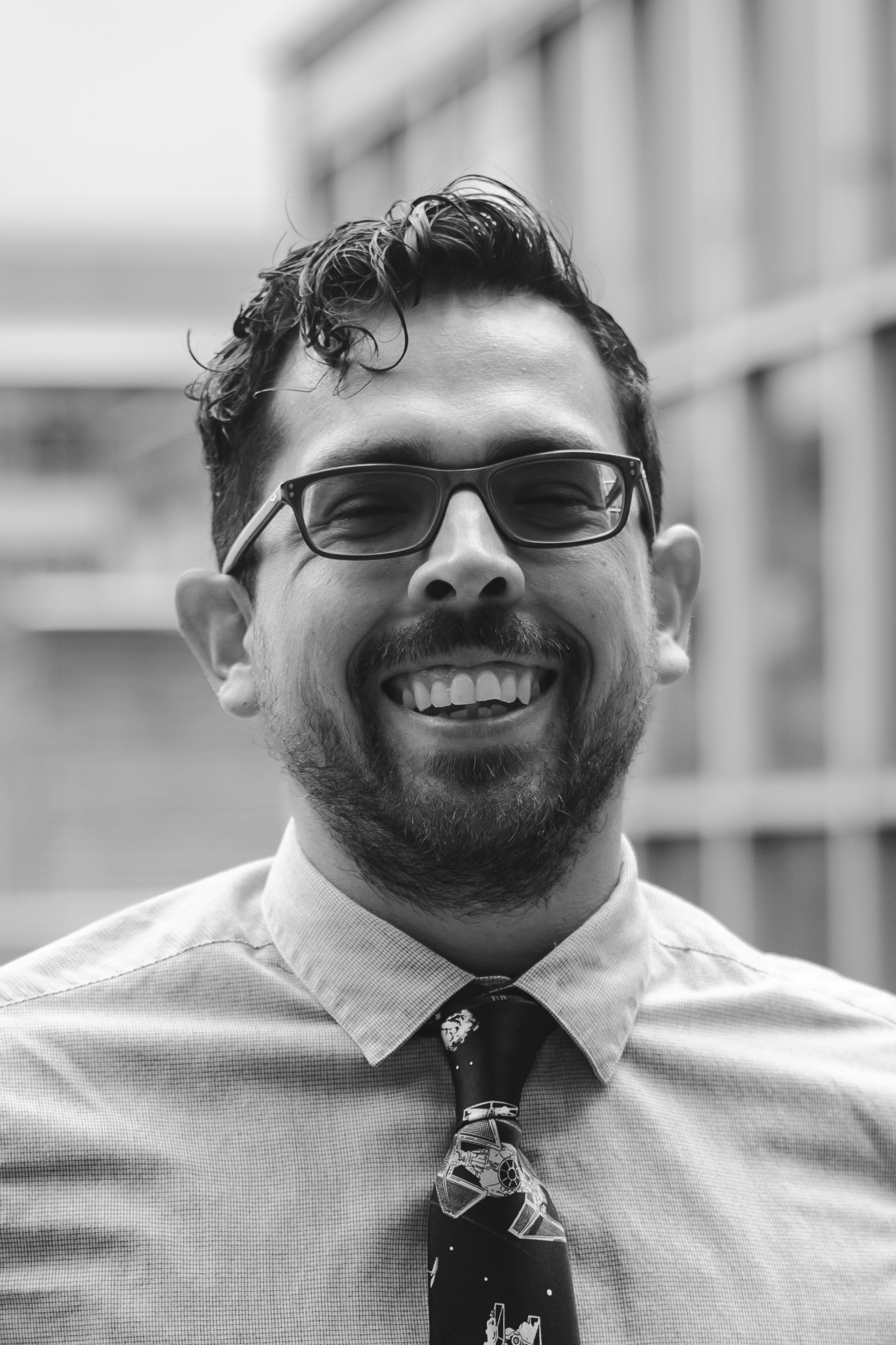 Black and white portrait of a smiling man with glasses, a beard, and messy hair, wearing a collared shirt and a Star Wars-themed tie, standing outdoors with blurred buildings in the background.