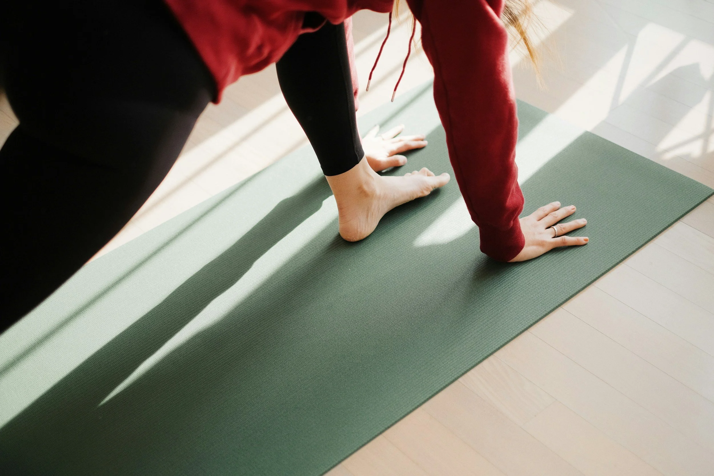 Person doing a yoga pose on a green mat indoors in sunlight, wearing a black shirt and red sweatshirt.