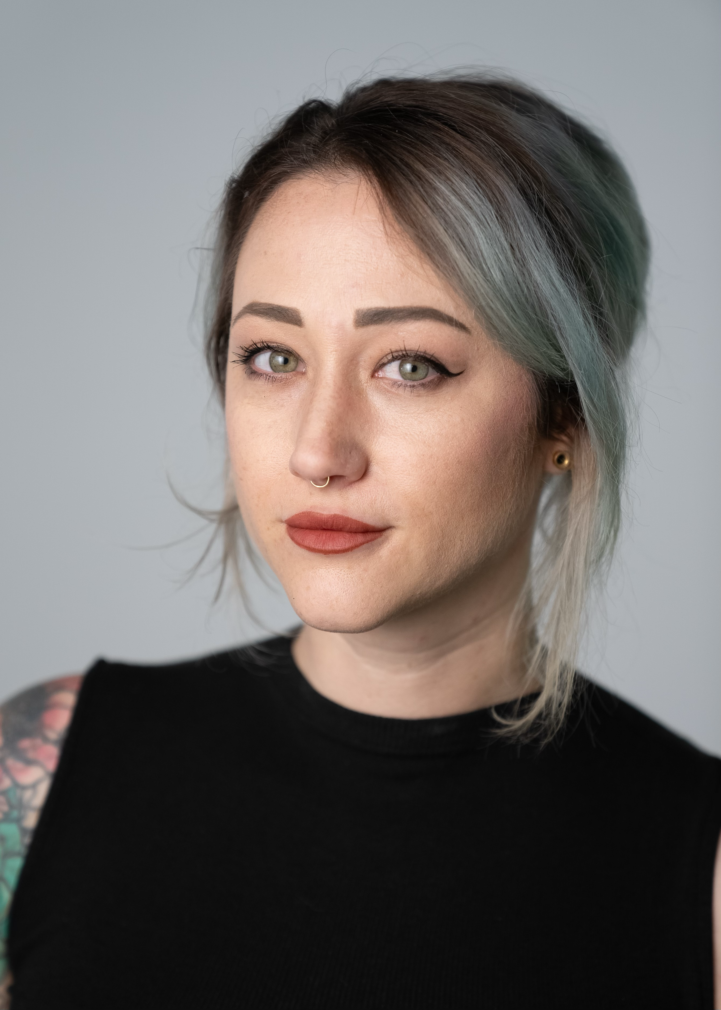 Close-up of a woman with short dark hair with gray streaks, green eyes, arched eyebrows, winged eyeliner, septum piercing, wearing makeup with a matte lipstick, black sleeveless top, and tattooed arm, against a plain gray background.