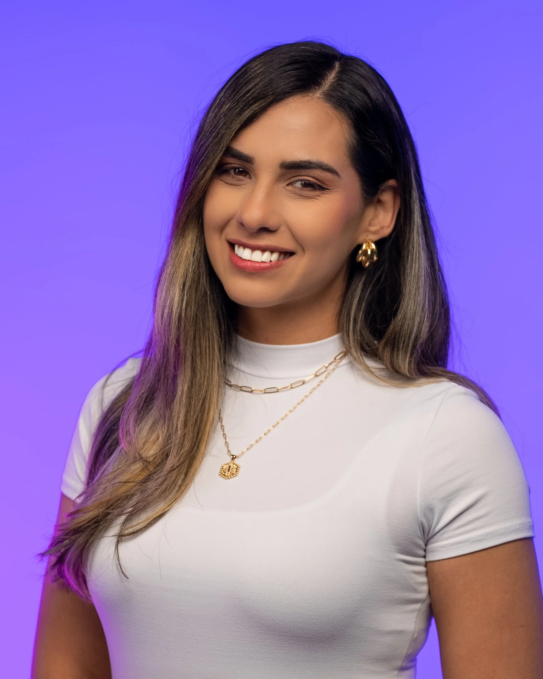 A woman with long, wavy, brown and blonde hair, wearing gold earrings, a white t-shirt, and layered gold necklaces, smiling against a purple background.