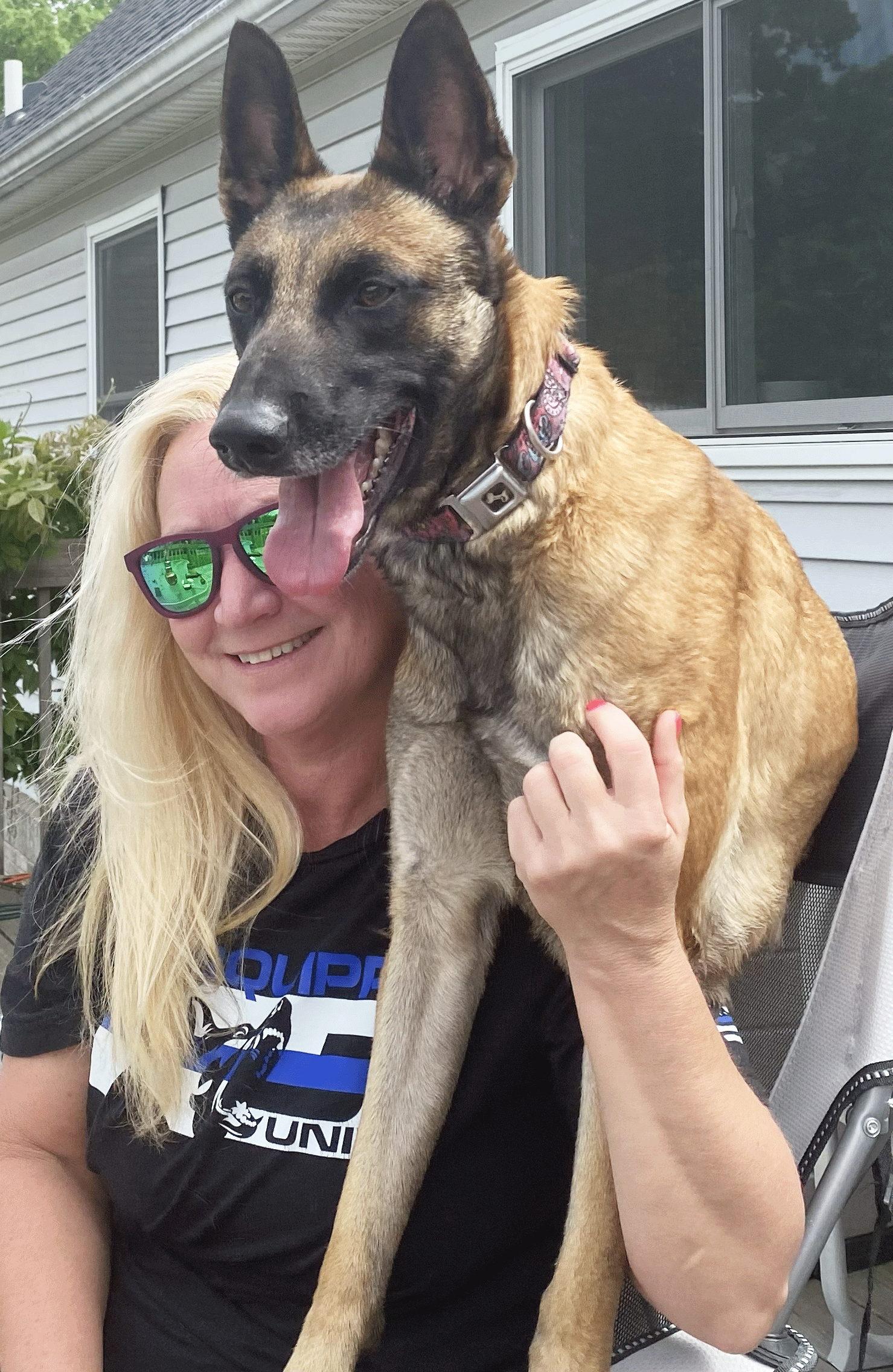 A woman with blonde hair, wearing sunglasses, smiling, holding a large Belgian Malinois dog with erect ears and tongue hanging out, outside near a house with white siding.