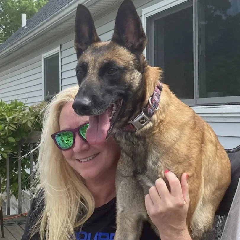 A woman with blonde hair and green sunglasses smiling while holding a large Belgian Malinois dog outside near a house with white siding.