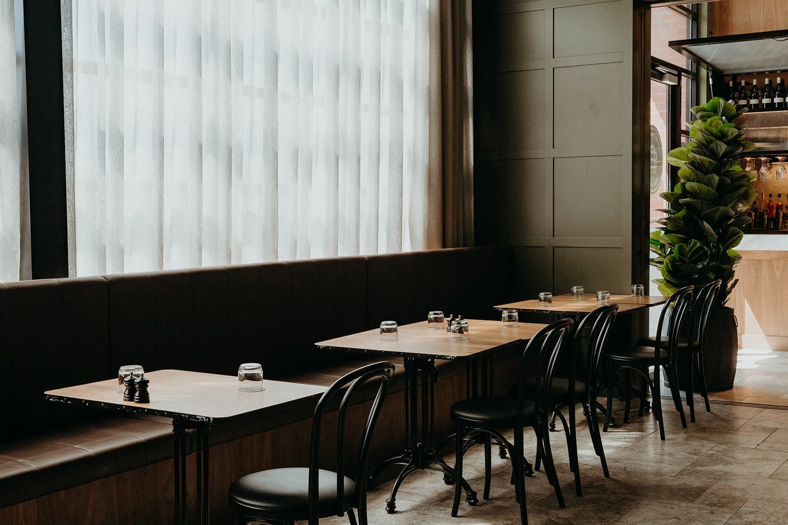 Empty restaurant with wooden tables set with glasses, black chairs, a long black leather bench seat, large window with sheer curtains, green plant, and a bar area in the background.
