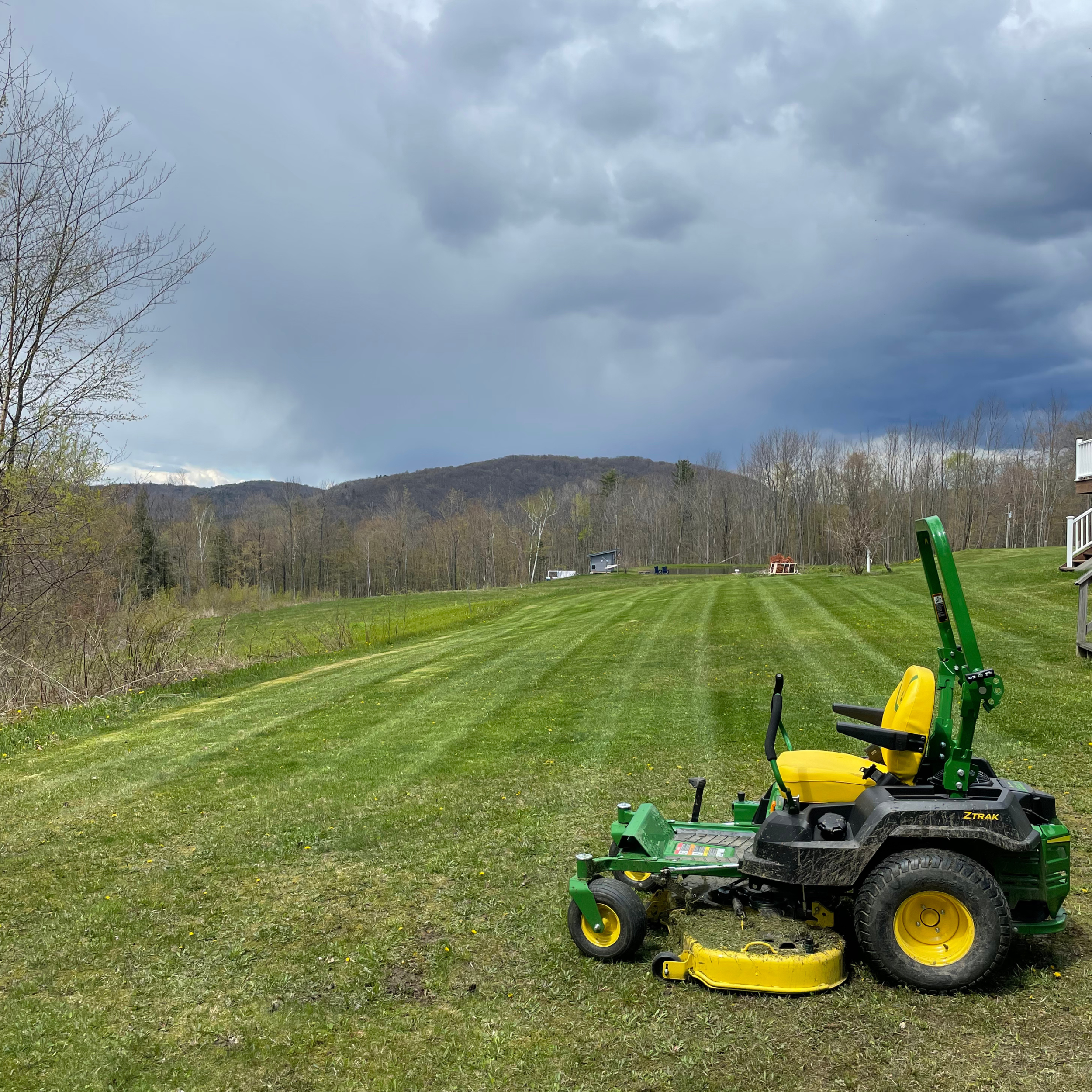 A green and yellow riding lawn mower on a freshly mowed grassy yard with a backdrop of trees, hills, and a cloudy sky.