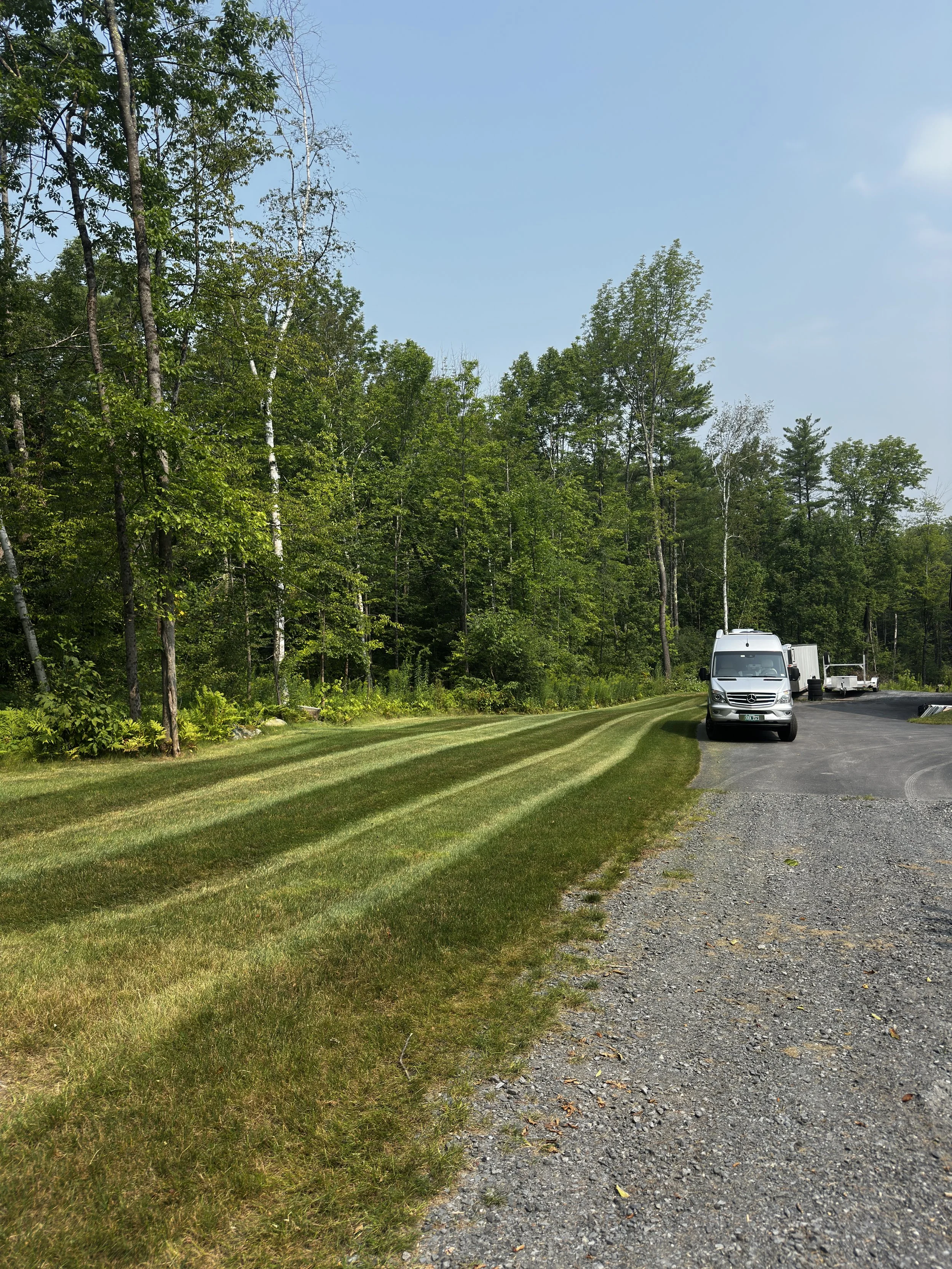 A gravel driveway with a row of freshly mowed grass on the side, leading into a wooded area with tall green trees under a clear blue sky, and a silver van parked on the driveway.