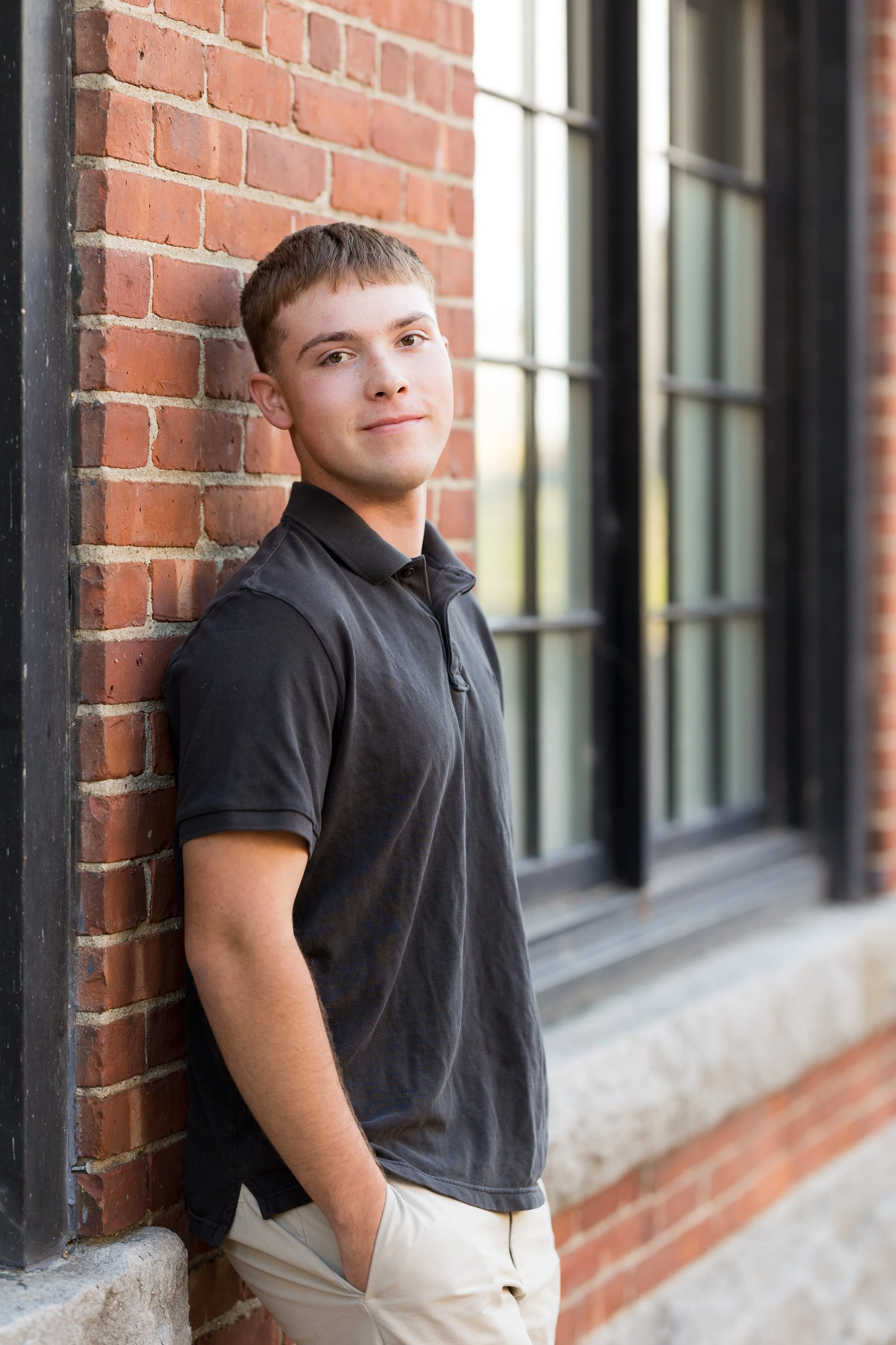 A young man in a black polo shirt and beige pants leaning against a red brick wall with large black-framed windows behind him.