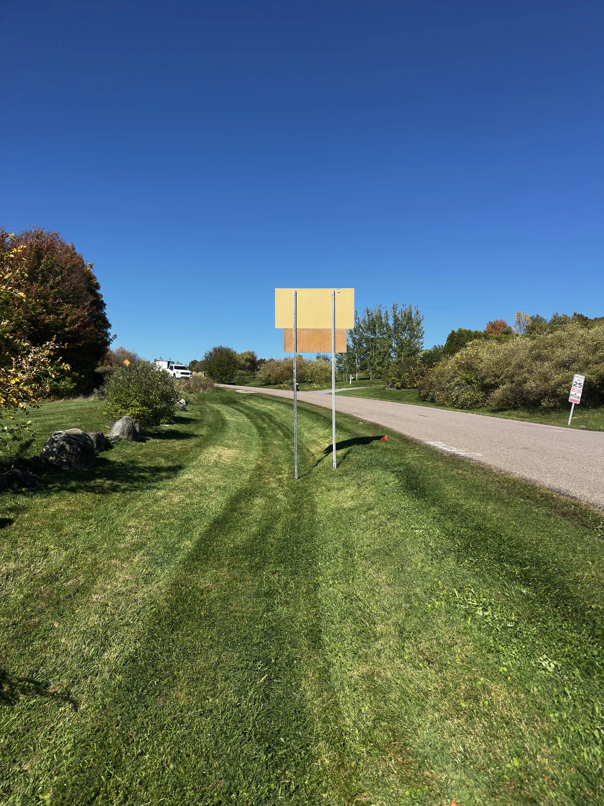 Empty billboard sign along a grassy roadside with trees, bushes, and a clear blue sky in the background.