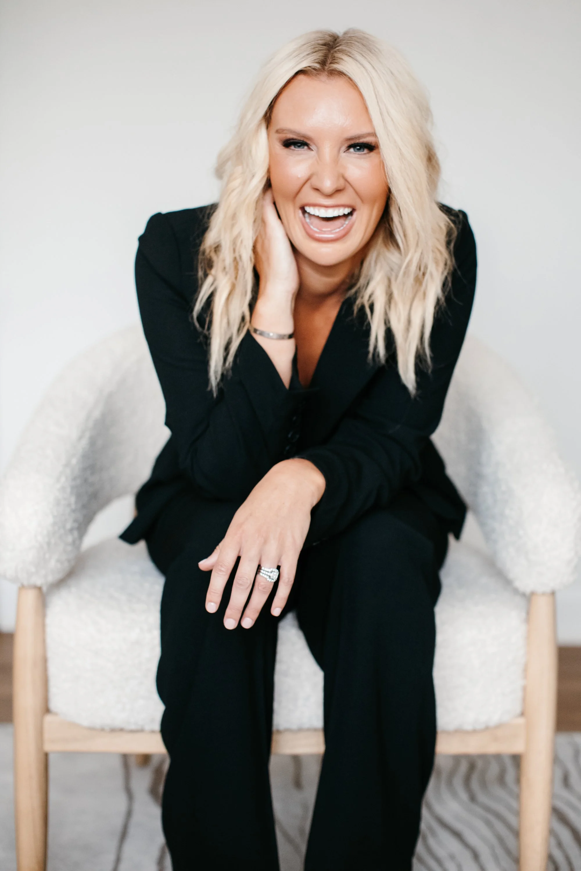 A woman with blonde hair, smiling and laughing, sitting on a white, textured chair against a plain background, wearing a black blazer and black pants.