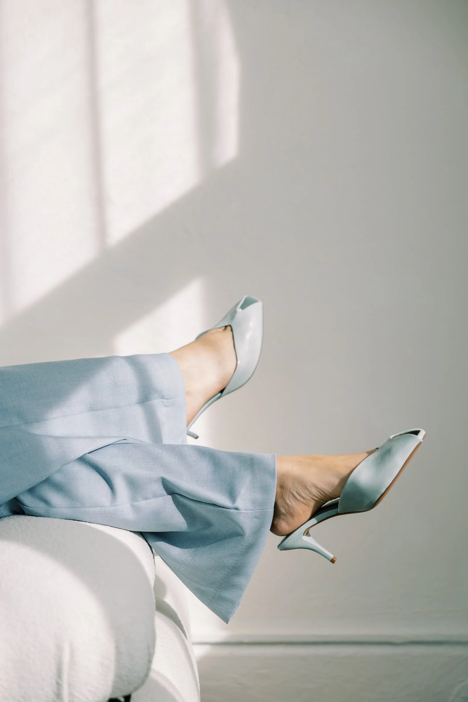 Woman’s legs in light blue pants and beige high heels, resting on furniture with a minimalist background and shadows on the wall.