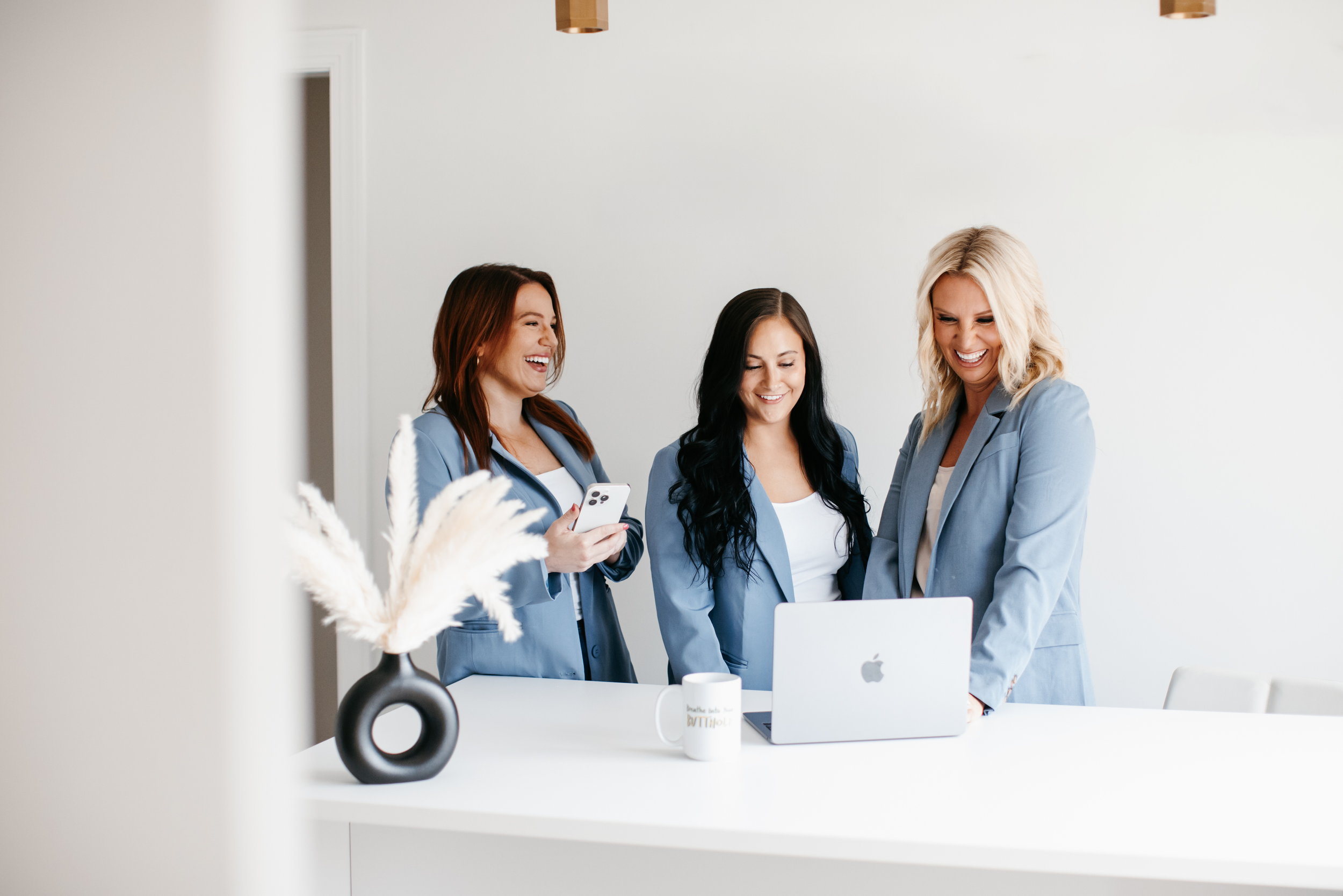 Three women in blue blazers standing around a white table, looking at a laptop and smiling, with a coffee mug and a vase with white dried flowers on the table.