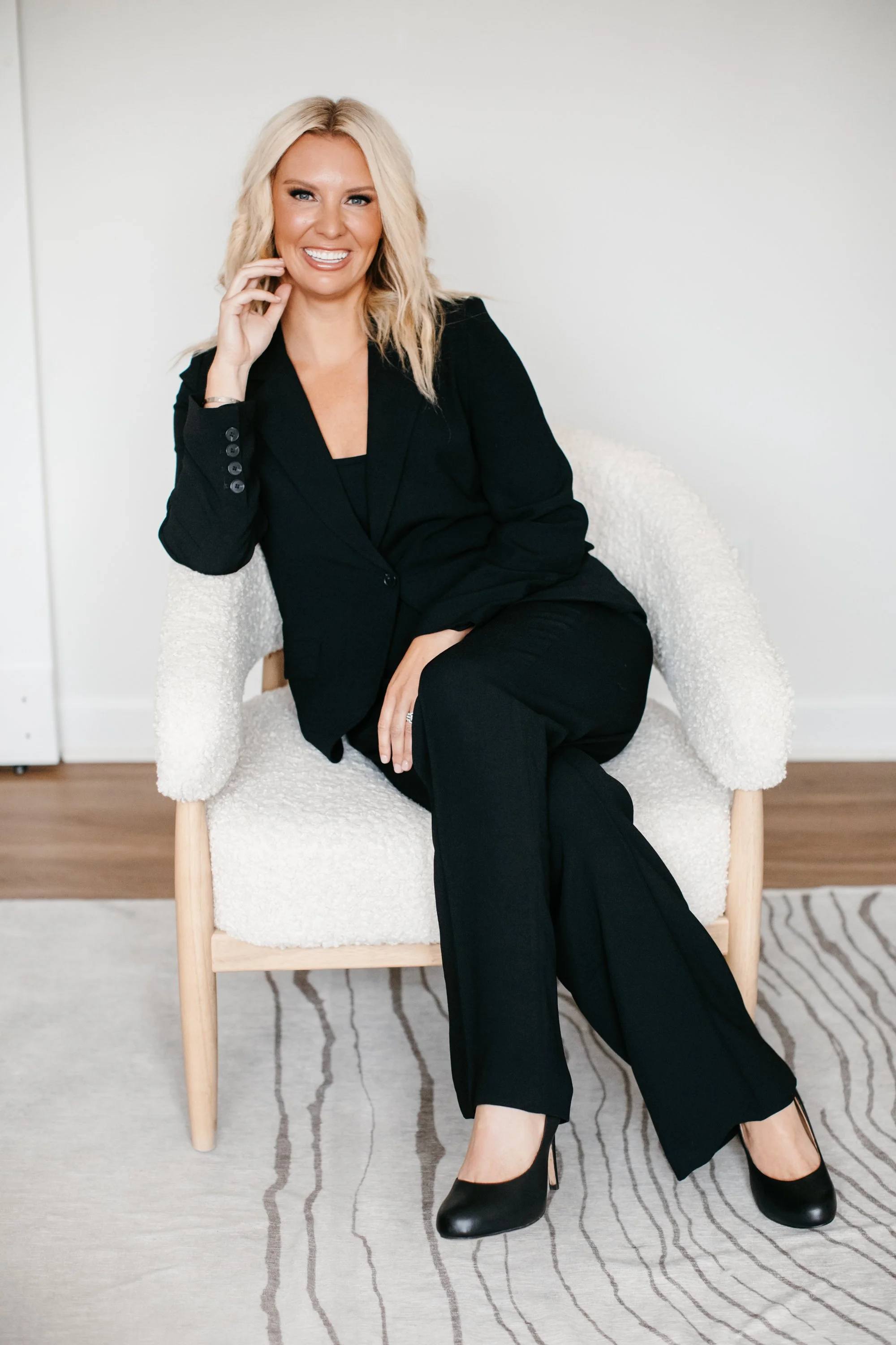 Dr. Audra Lance in a black suit sitting on a white textured chair, smiling, with her legs crossed, in a minimalistic room.