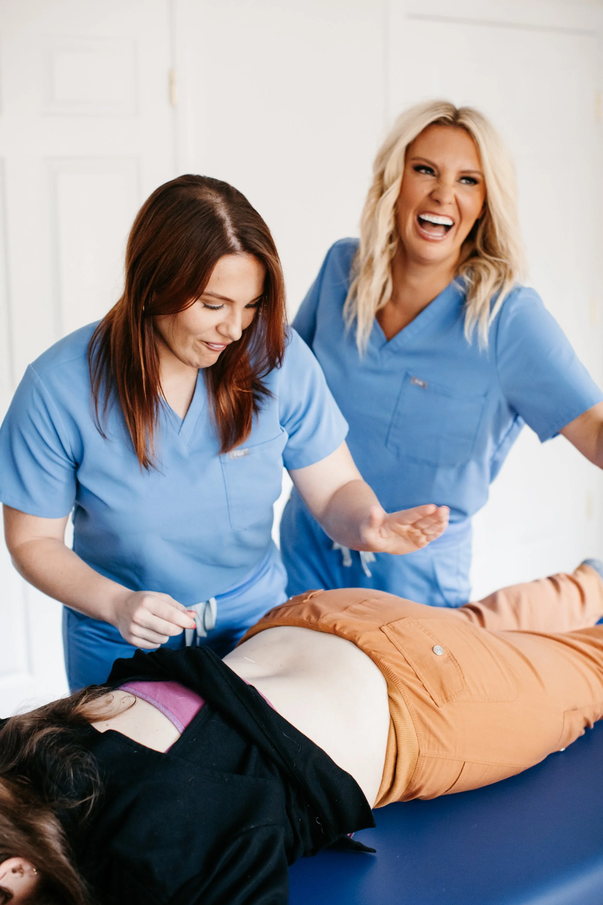 Two female healthcare workers in blue scrubs appear to be doing chiropractic work on a woman lying on a table, who is wearing an orange pair of pants and a black top, during a training session or simulation.