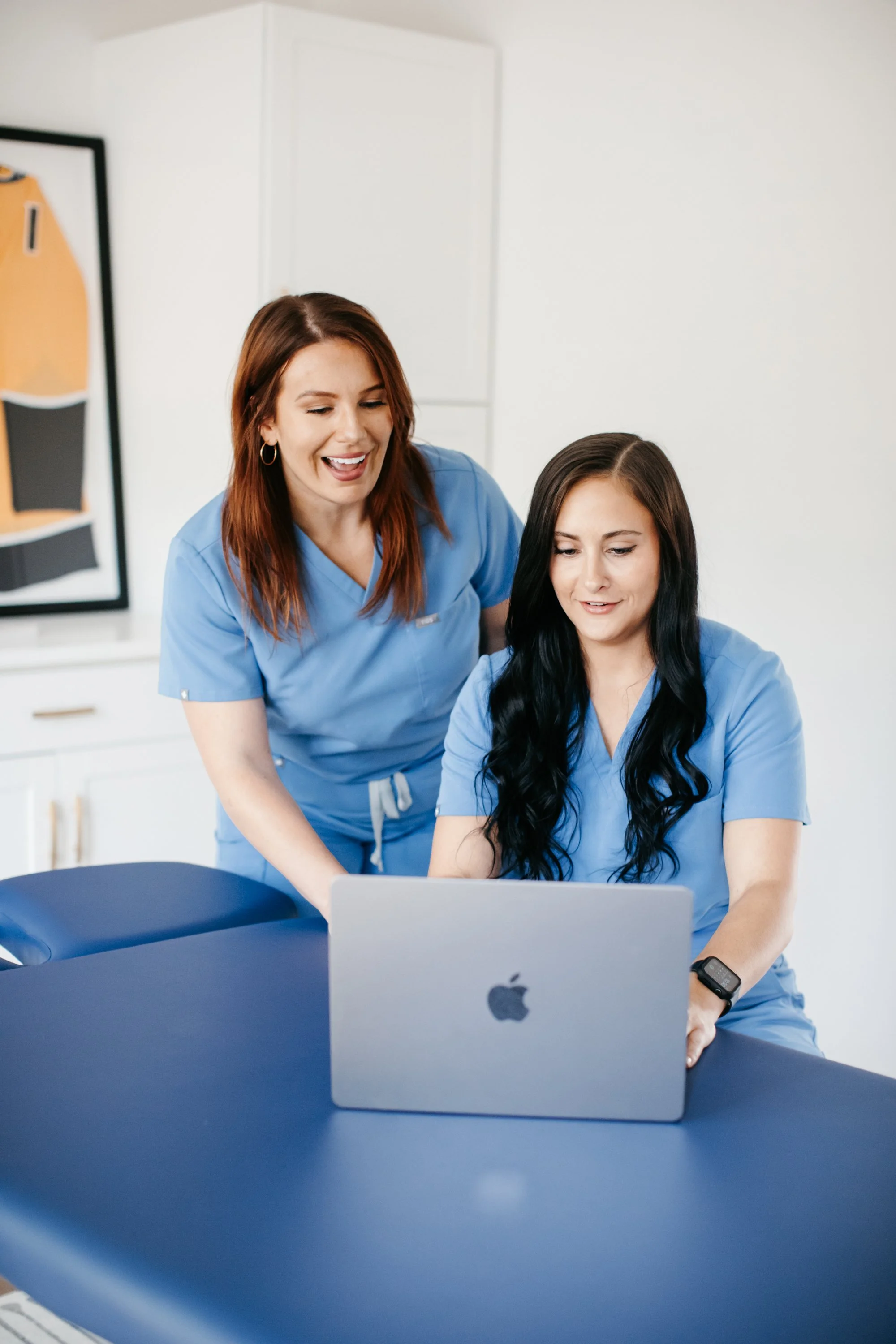 Two women in blue scrubs looking at a laptop, one sitting at a table and the other standing beside her, in a medical setting.