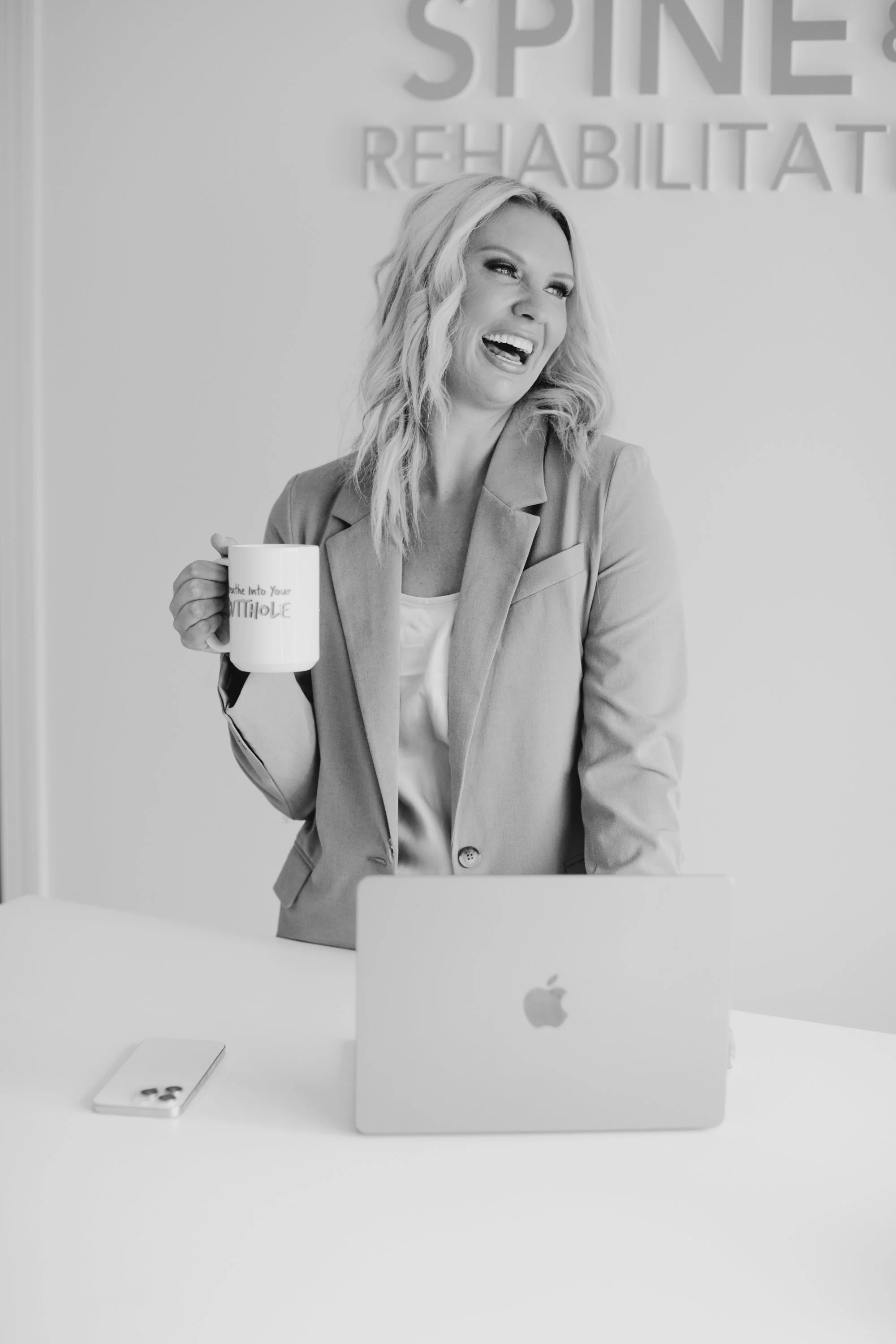 Dr. Audra Lance laughing while holding a coffee mug, standing behind a desk with a laptop and smartphone, in an office.