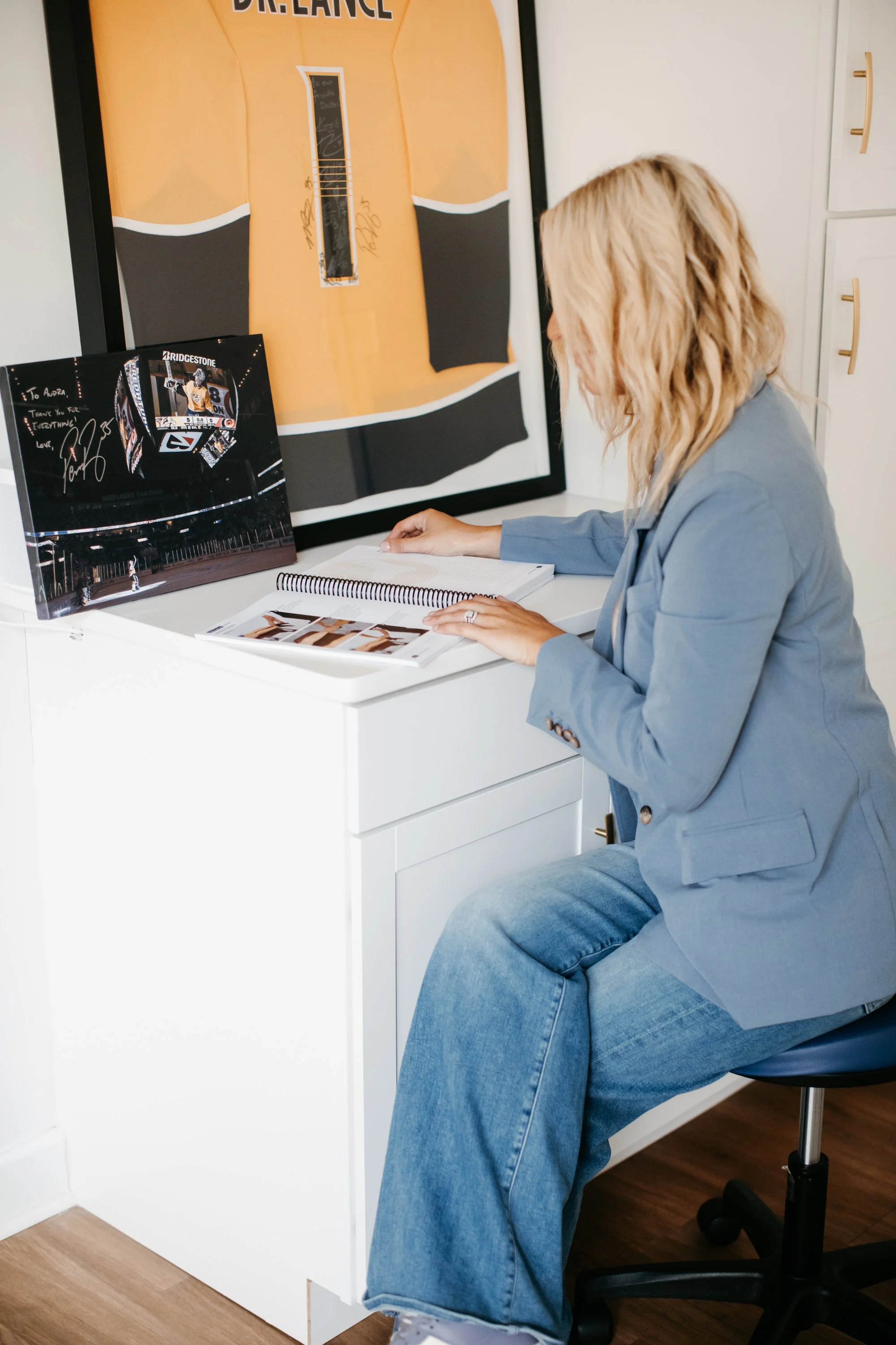 Dr. Audra Lance sitting at a white desk, reading a book, with a framed signed hockey puck and a large framed hockey jersey hanging on the wall behind her.