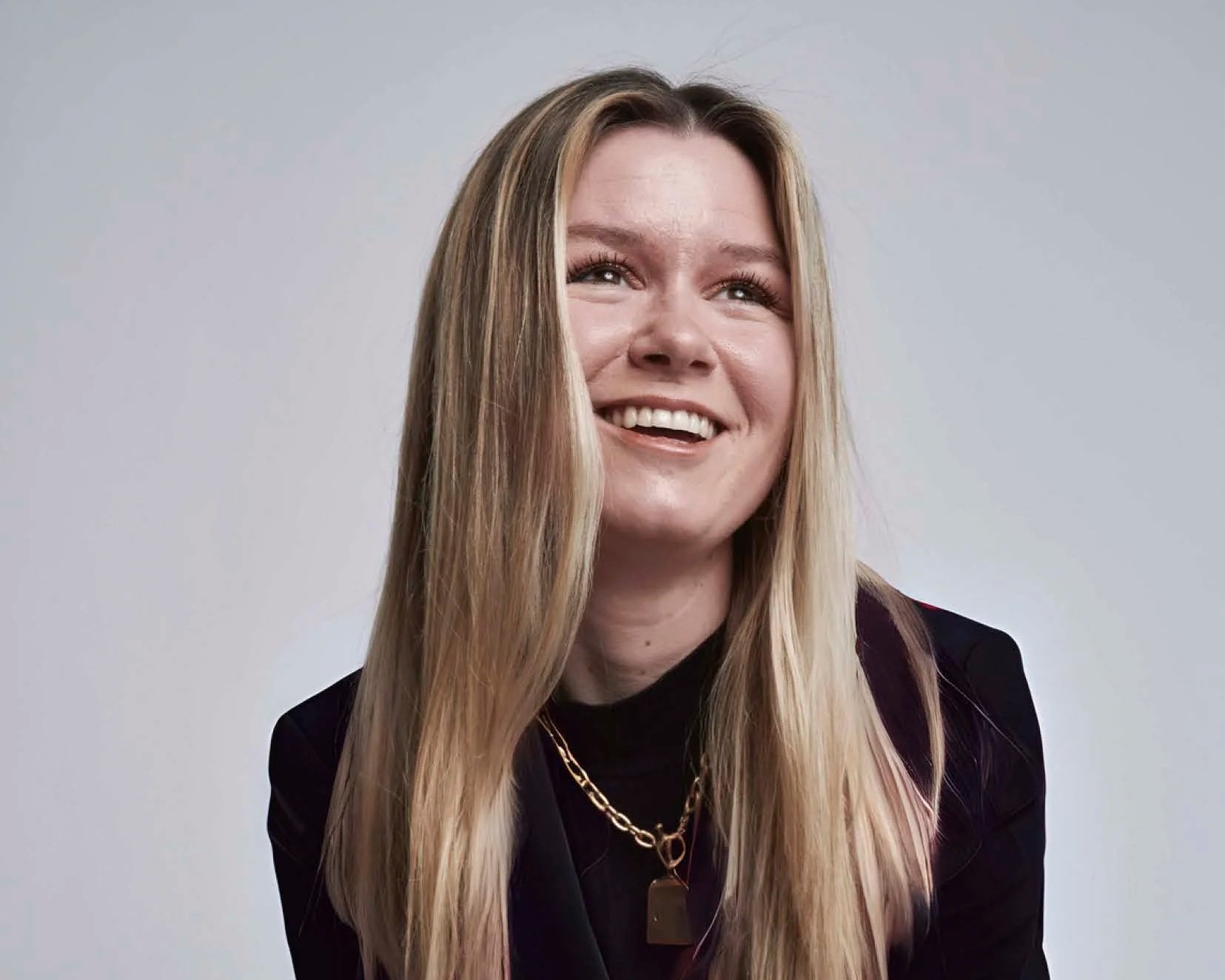 A woman with long blonde hair smiling, wearing a black top, gold chain necklaces, and a black blazer, against a plain light gray background.
