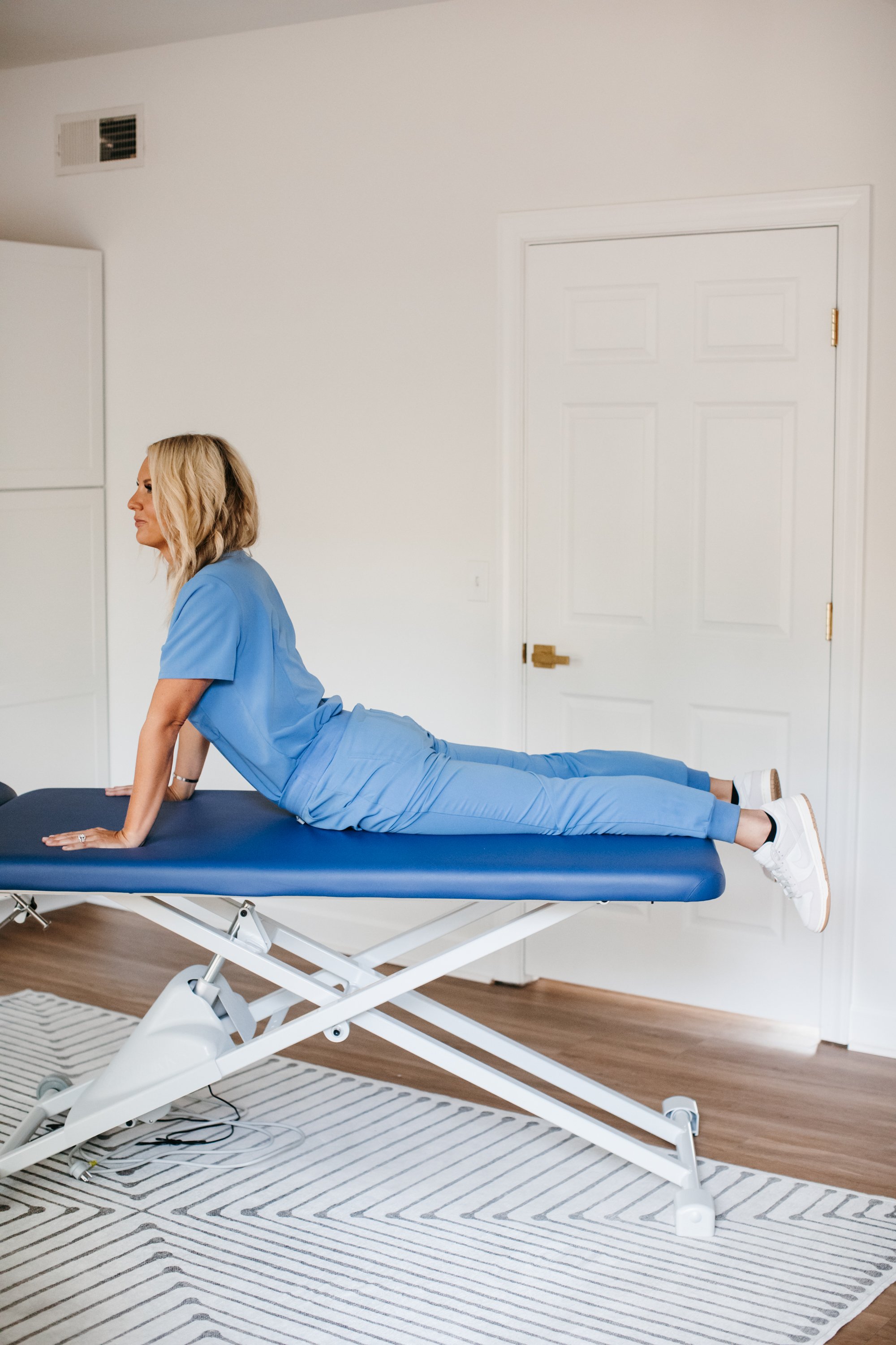 A woman in blue medical scrubs performing the Cobra pose on a medical examination table in a room with white walls, wood flooring, a cabinet, and a closed white door.