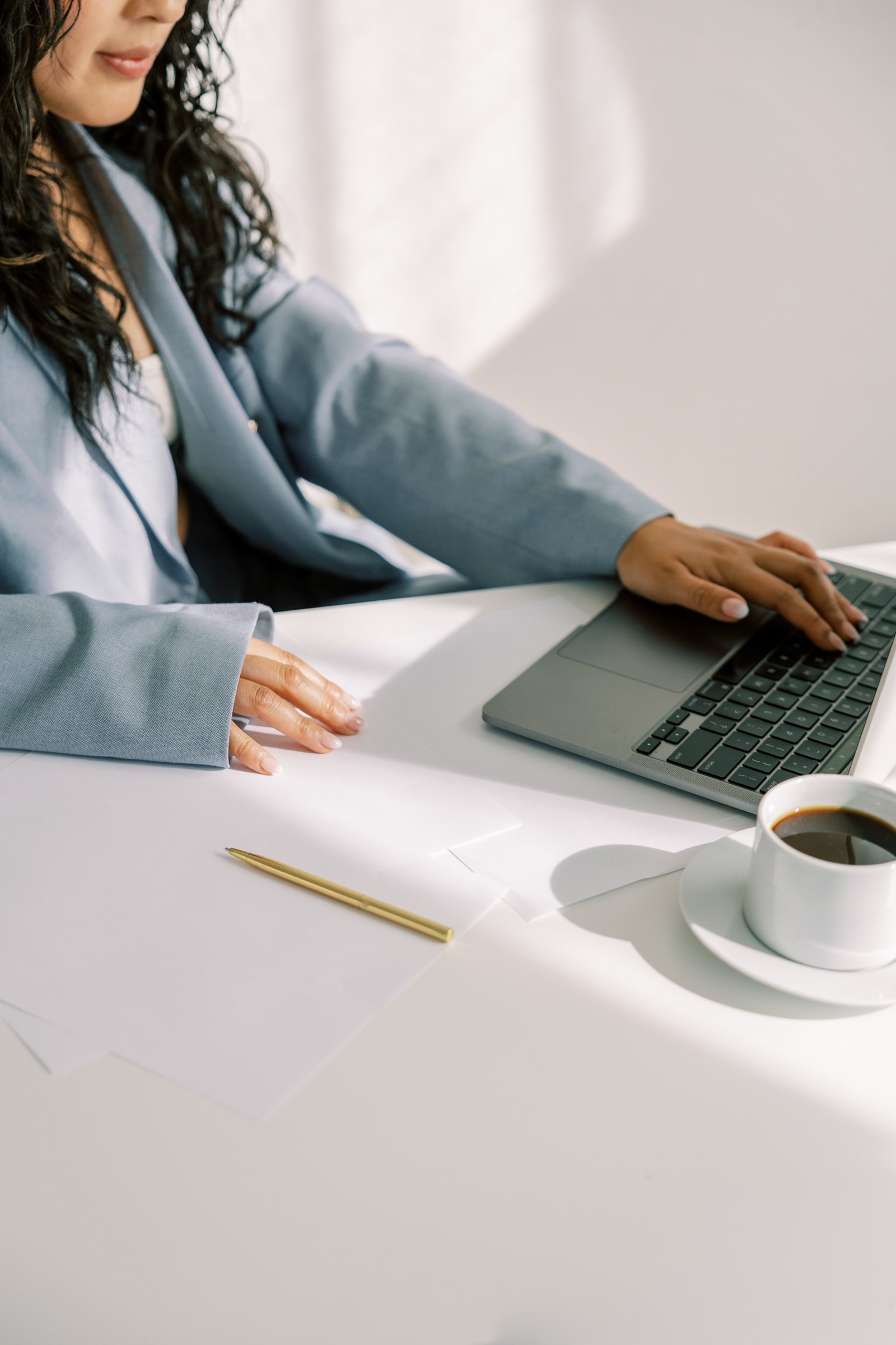 A person sitting at a white desk working on a laptop, with papers, a gold pen, and a cup of coffee nearby.