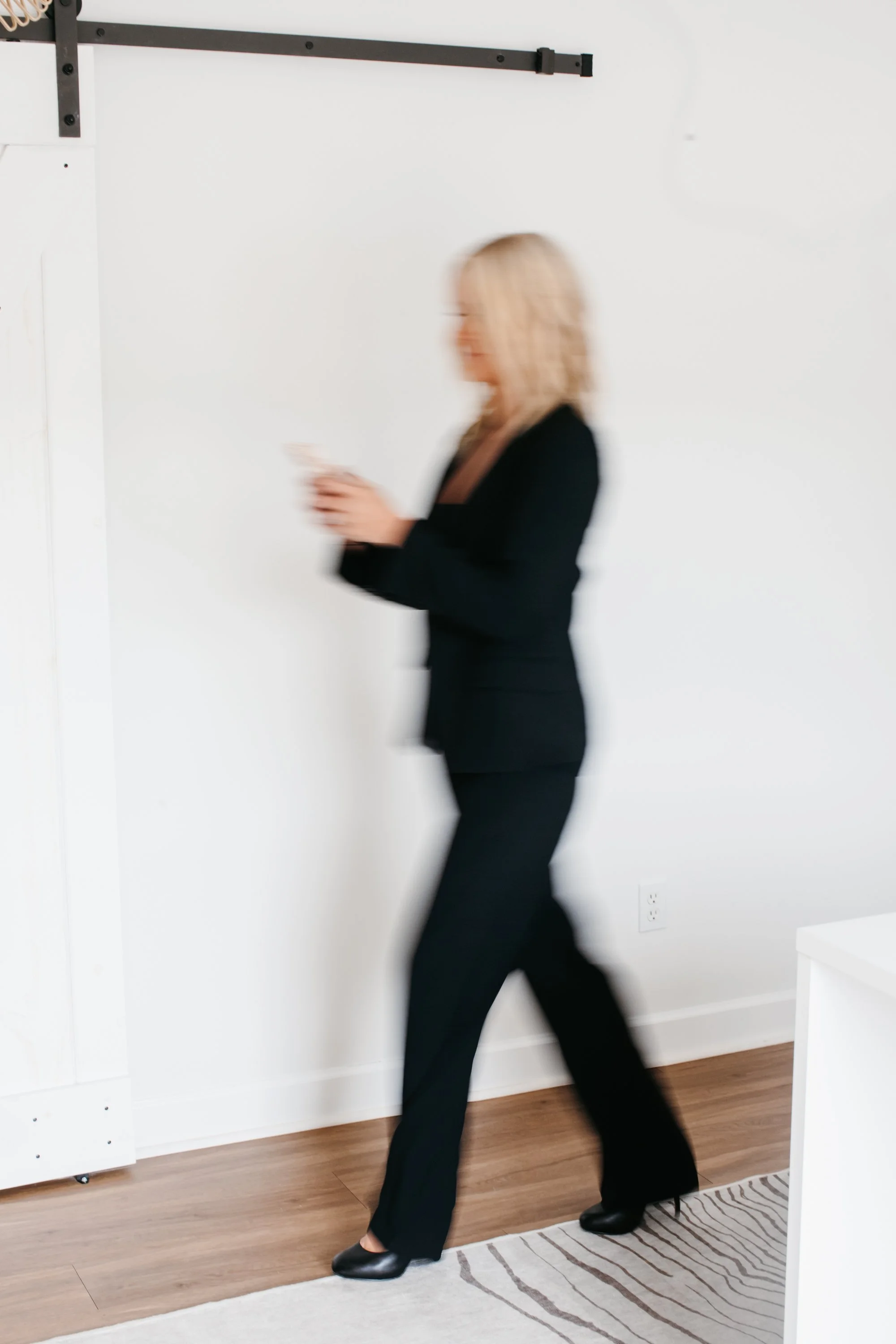 Blurry image of a woman in a black suit walking and looking at her phone in a white room with wooden flooring.