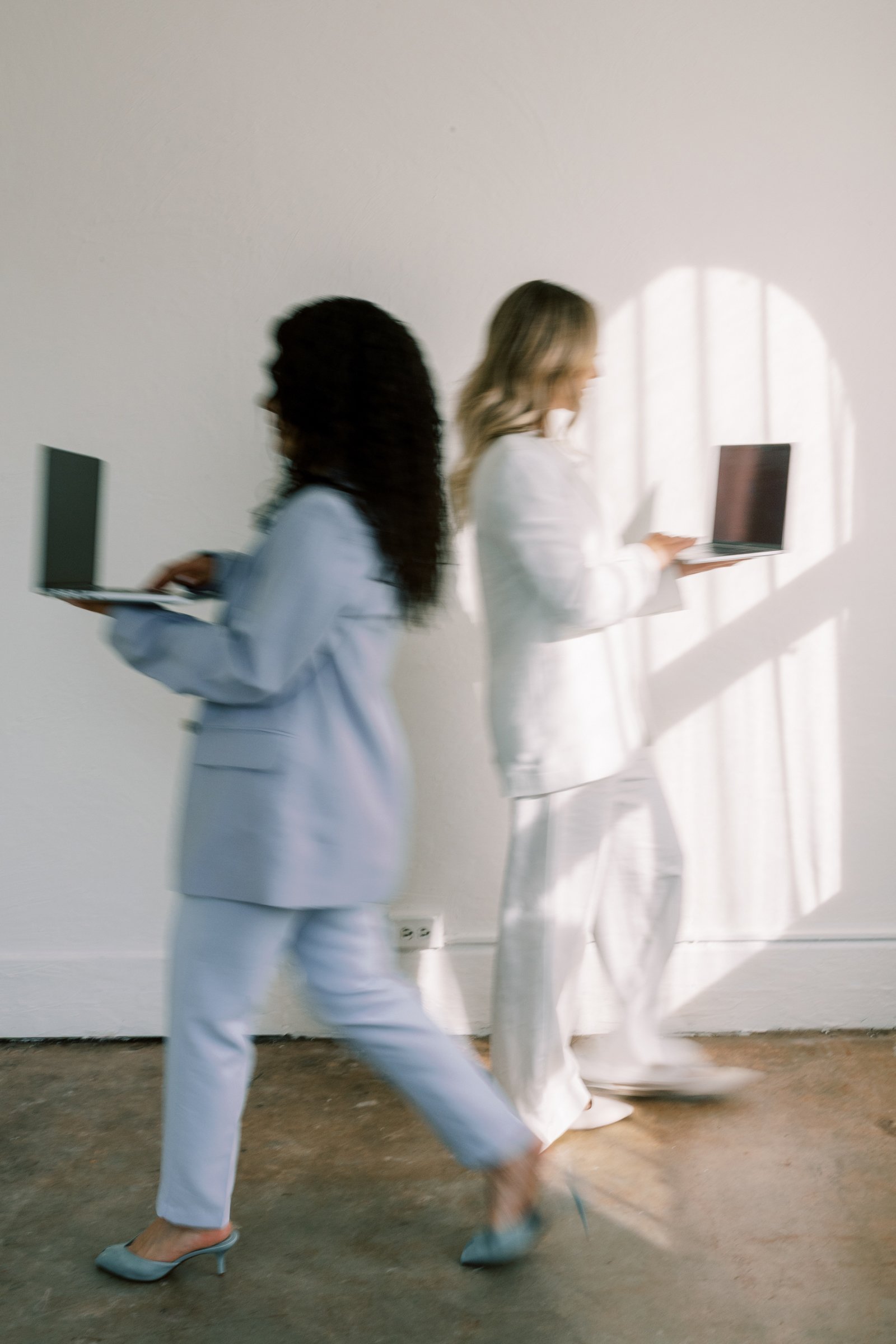 Two women walking side by side, each holding a laptop. They are dressed in white suits and heels, with sunlight casting shadows on the wall behind them.