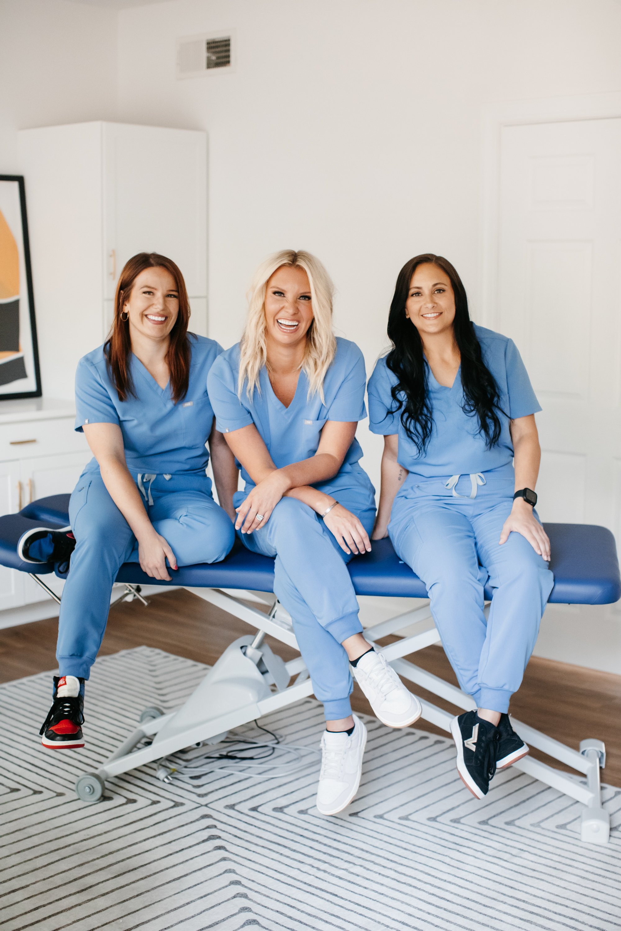 Three women in blue medical scrubs sitting on an examination table, smiling and laughing in a bright room.