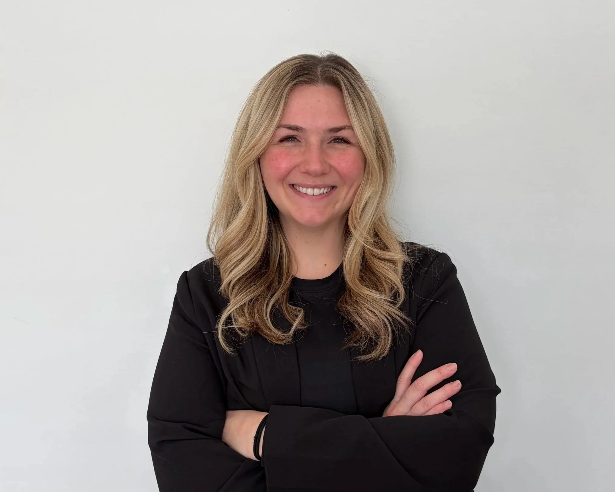 A woman with shoulder-length blonde hair, wearing a black blazer, smiling with arms crossed, standing against a plain white wall.
