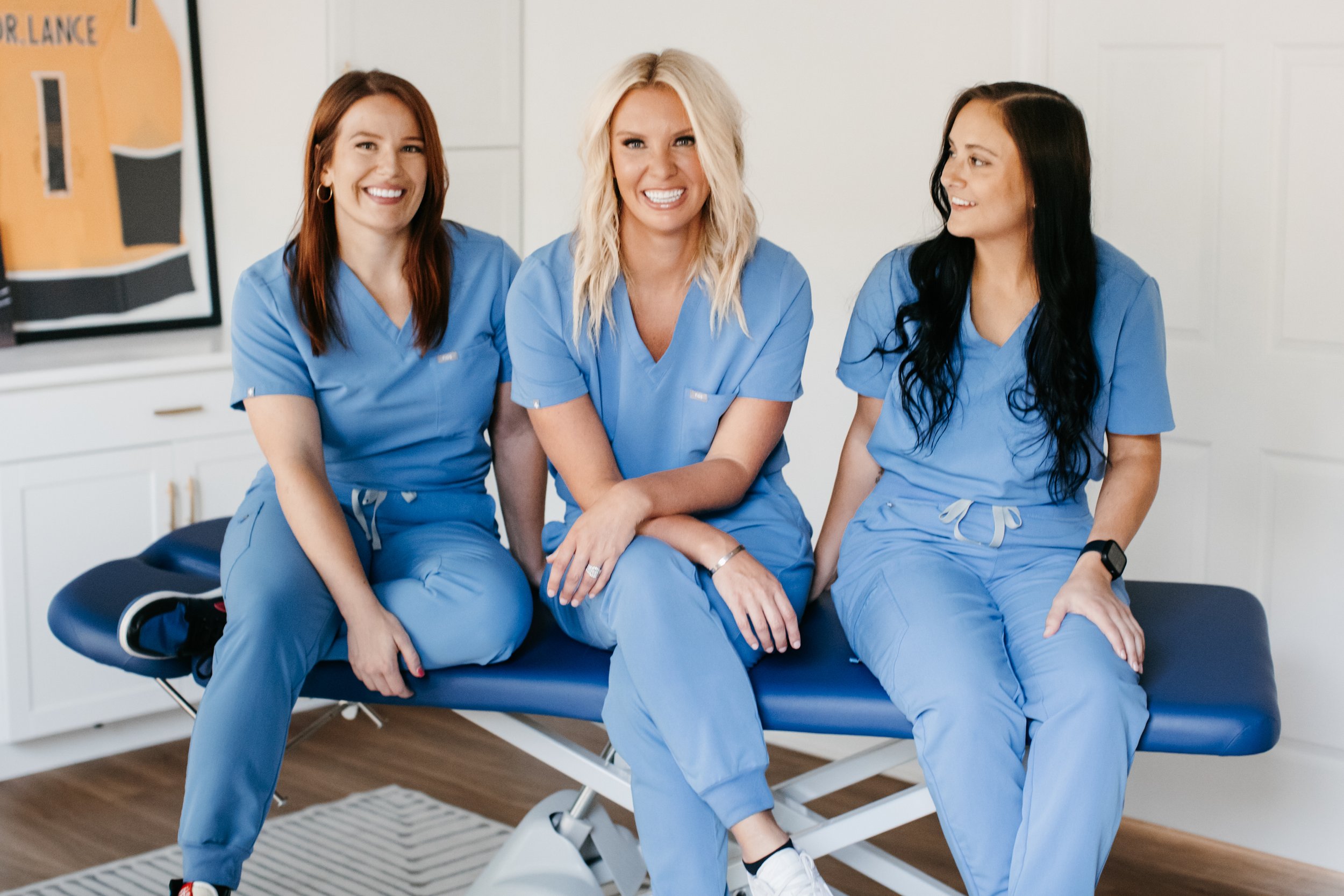 Three women in scrubs sitting on an examination table in a medical office, smiling and looking at each other.