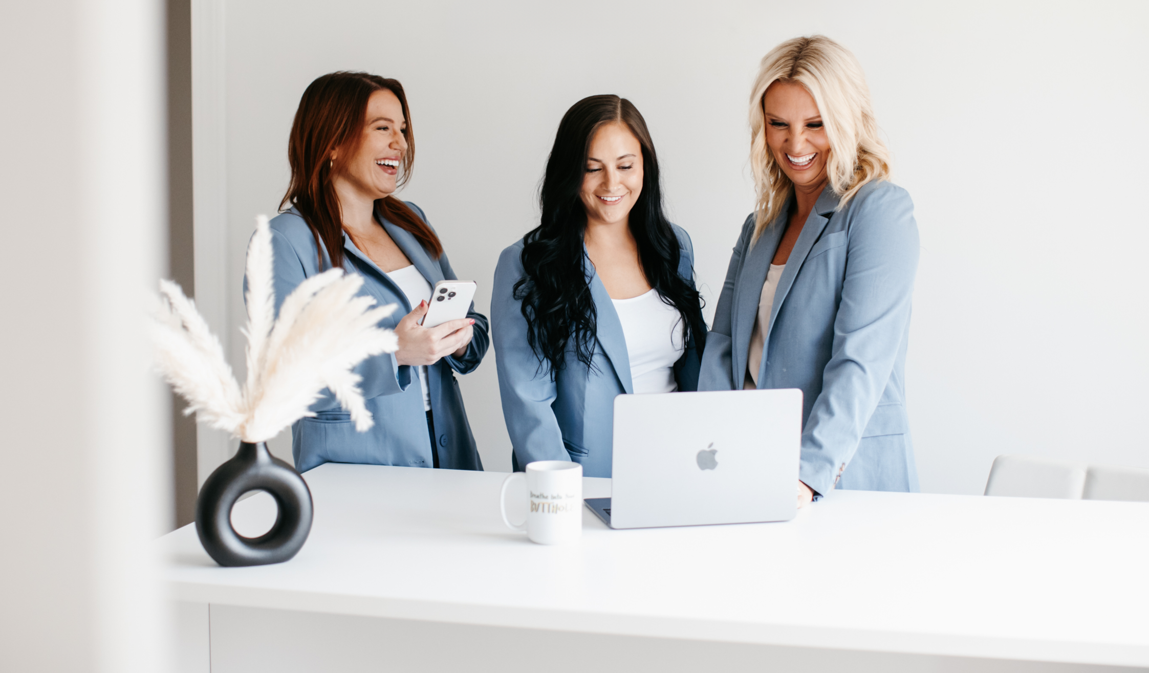 Three women in blue blazers gathered around a laptop in a modern office, smiling and engaging with each other.