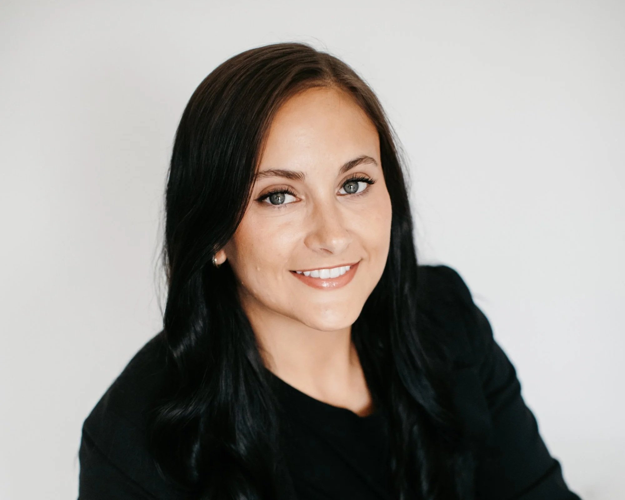 Close-up portrait of a woman with dark hair, blue eyes, wearing a black top, smiling against a plain light background.