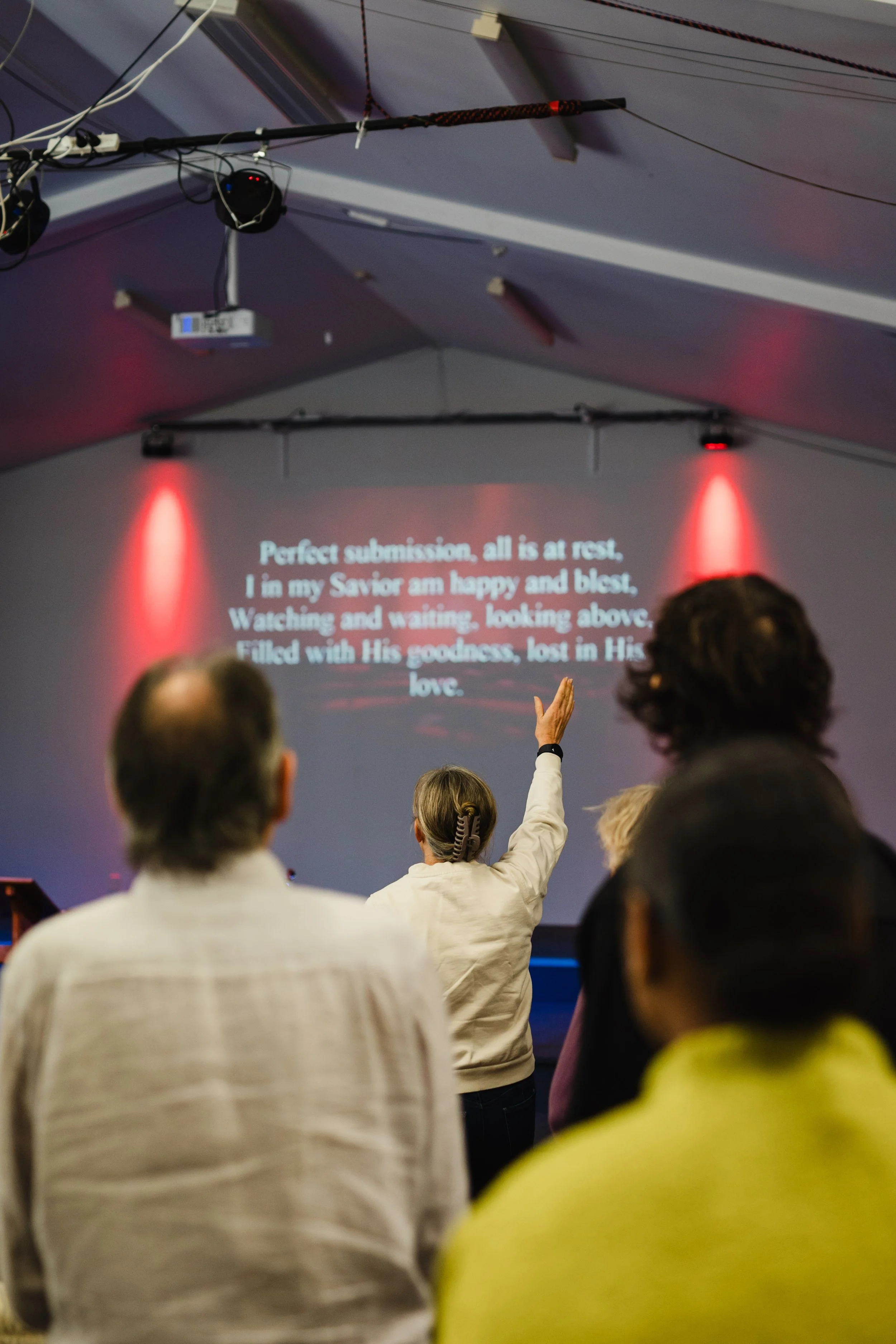 A group of people attending a church service or gathering in a large tent or hall, with one person raising their hand while a hymn or prayer is projected on the wall behind them.