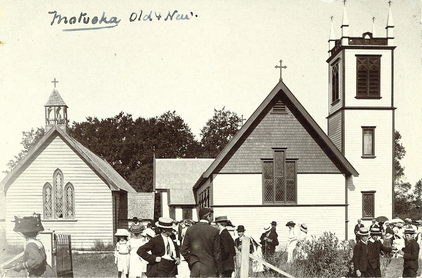 Historical black-and-white photo of a church with a tall steeple, surrounded by people dressed in early 20th-century clothing, outside in a yard with trees in the background, and handwritten note at the top reading 'Motueka Old & New'.