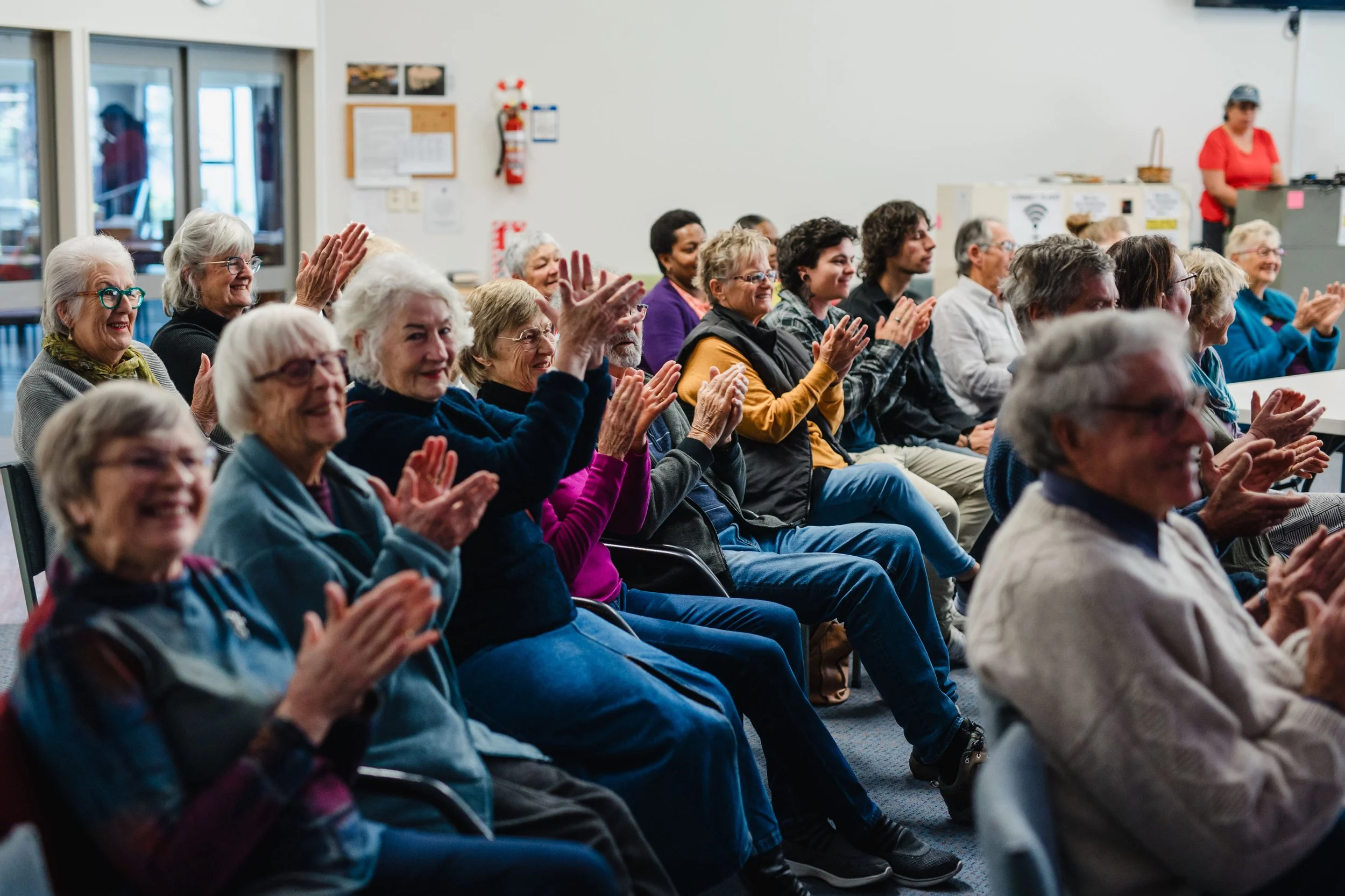 An audience of people clapping and smiling at an indoor event.