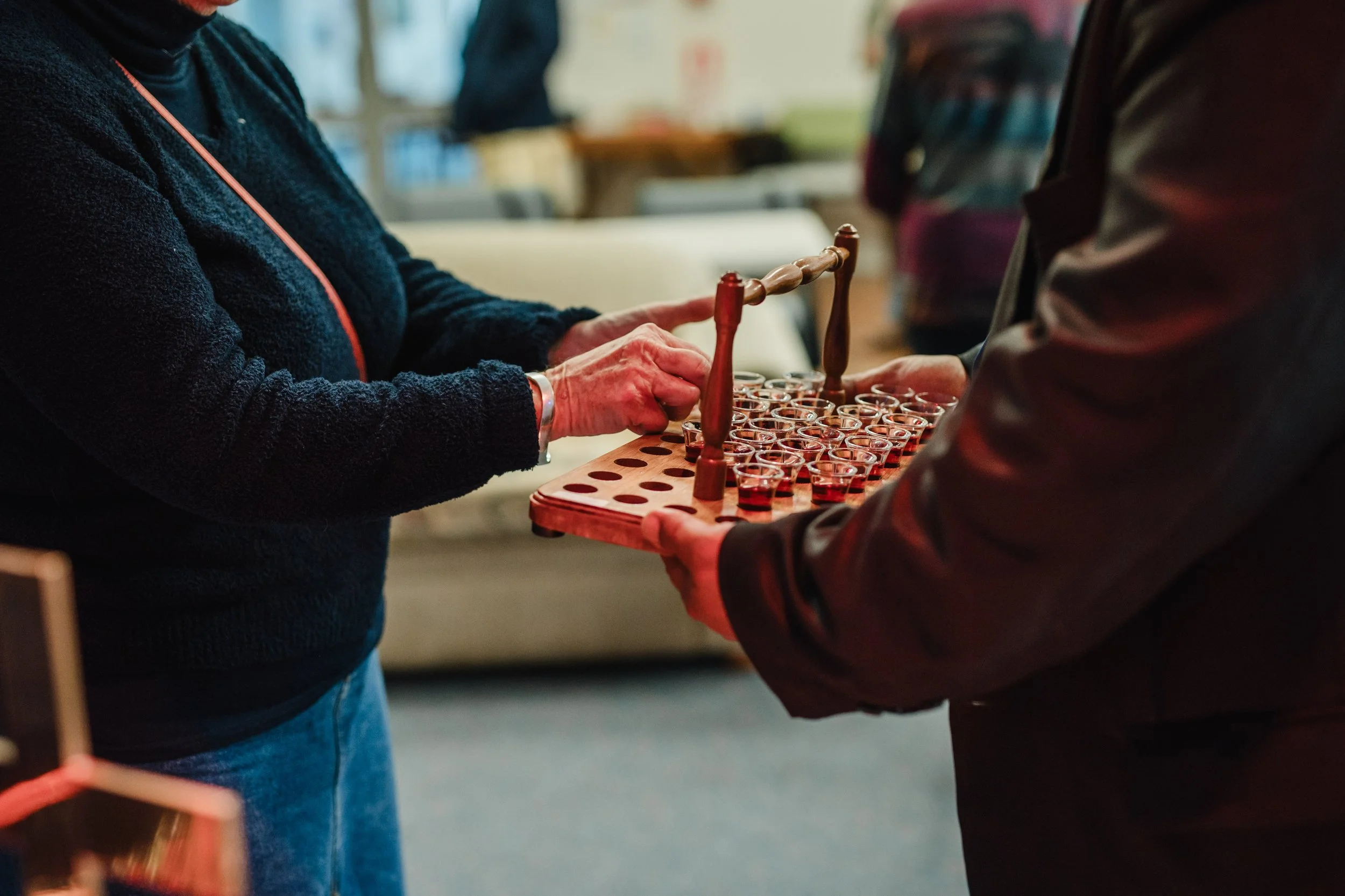 Two people are exchanging a wooden tray with small glasses filled with red liquid, possibly a tasting or sampling event.
