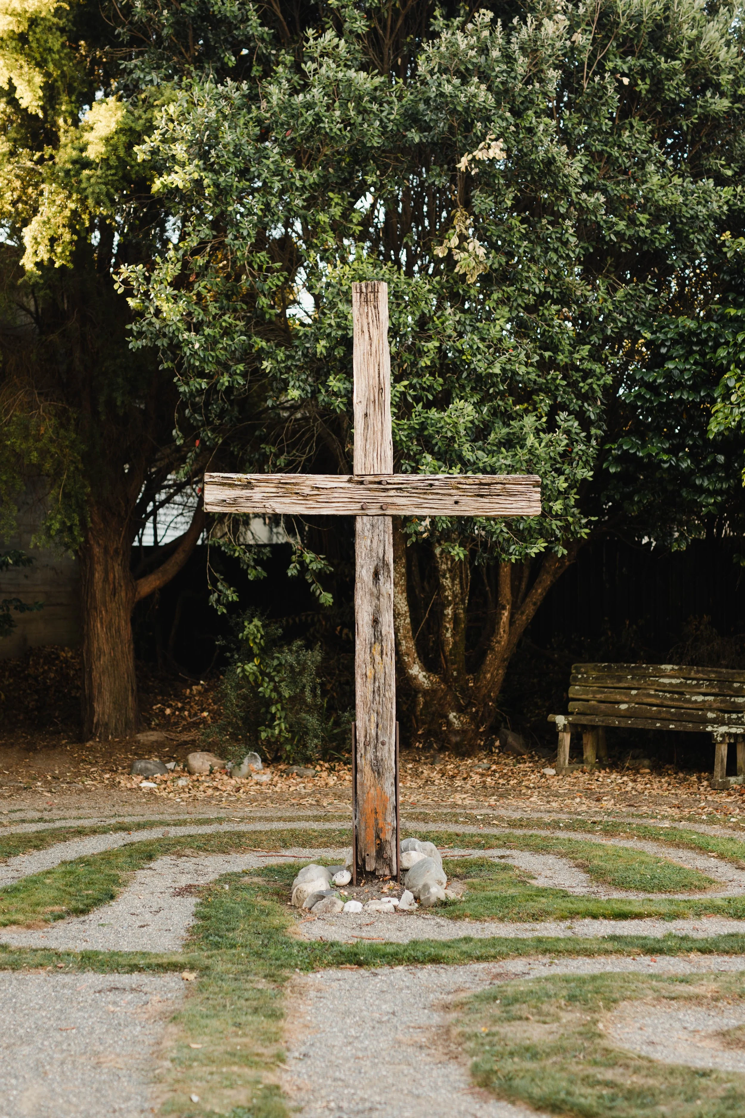 A weathered wooden cross situated in a landscaped garden with a gravel pathway and grass, surrounded by trees and a wooden bench in the background.
