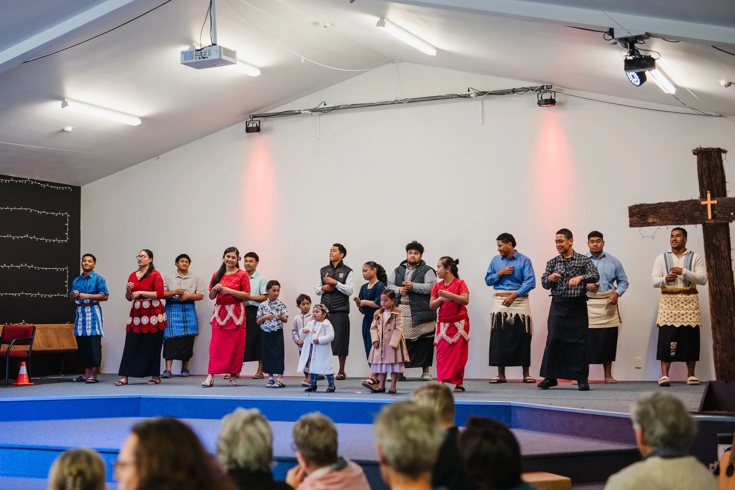 A group of people, including children and adults, wearing traditional clothing standing on a stage, with a large cross on the right side, in what appears to be a church setting.