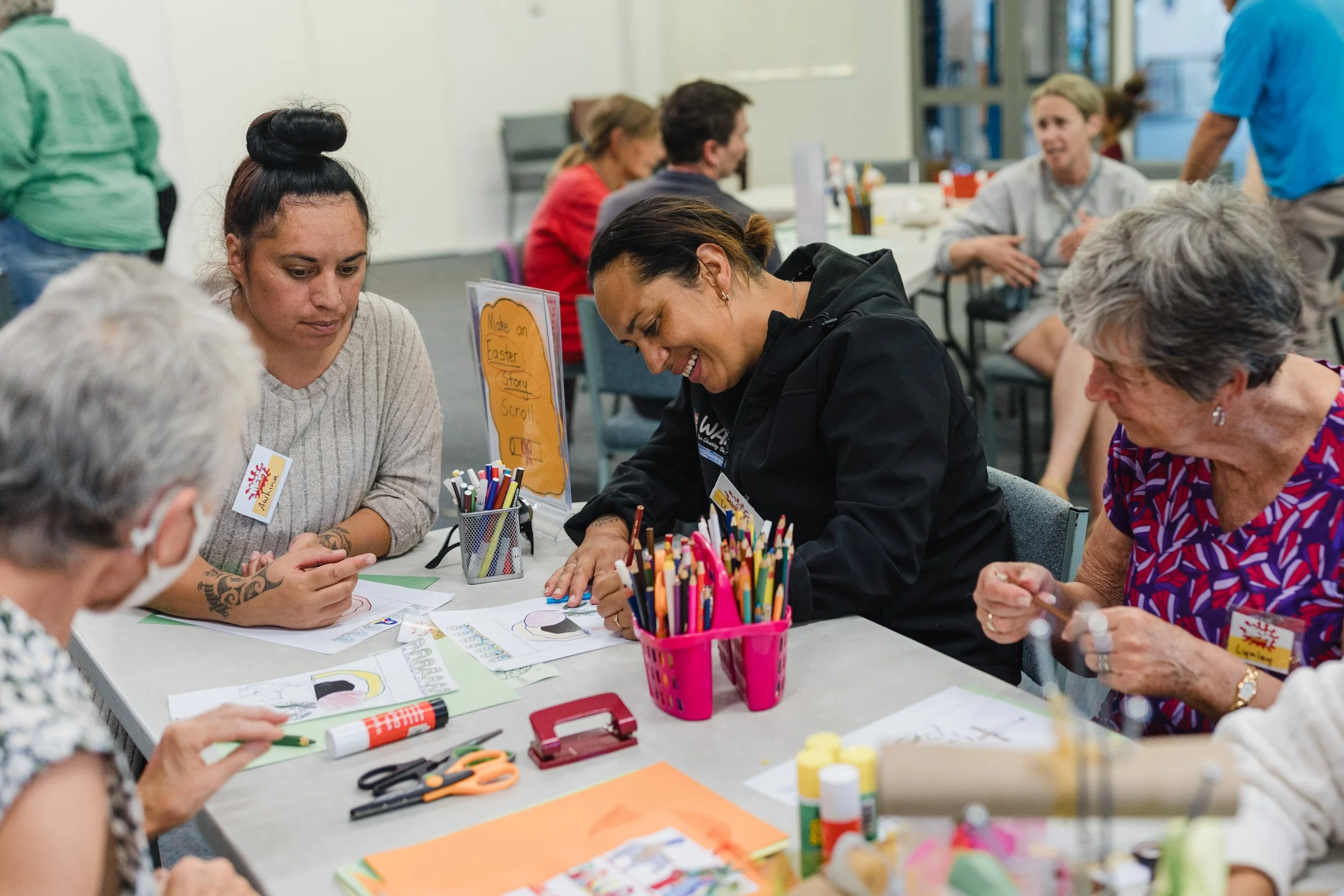 Group of women, including a young woman with a top bun hairstyle and tattoos, engaged in arts and crafts at a table with colored pencils, glue, scissors, and paper in a classroom or community center setting.