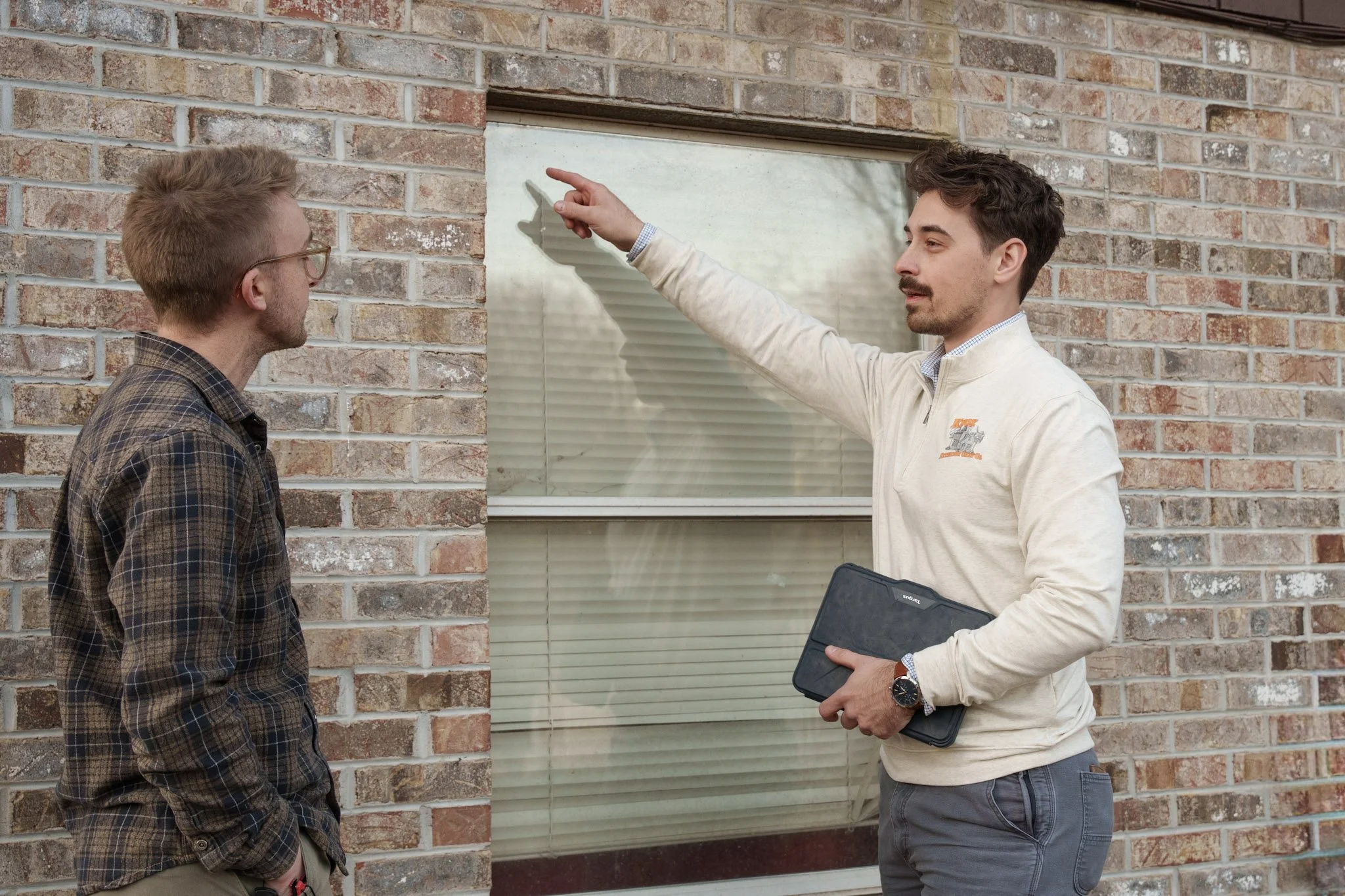 Two men having a conversation outside a brick building, one pointing at the window while the other listens.