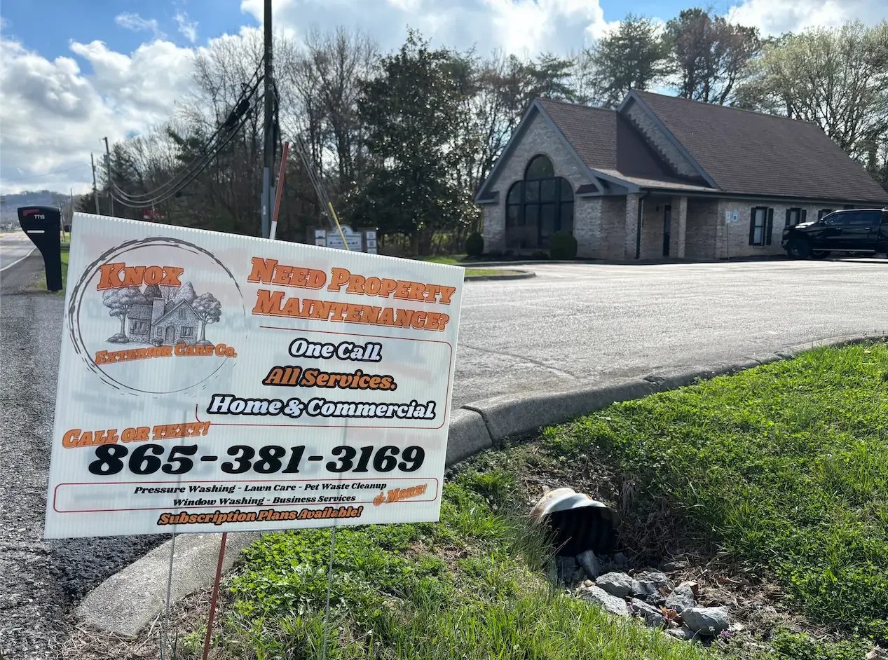 Sign advertising property maintenance services placed on a patch of grass next to a curb. In the background, there is a building with brick walls and a dark roof, cars parked outside, with trees and a cloudy sky behind.