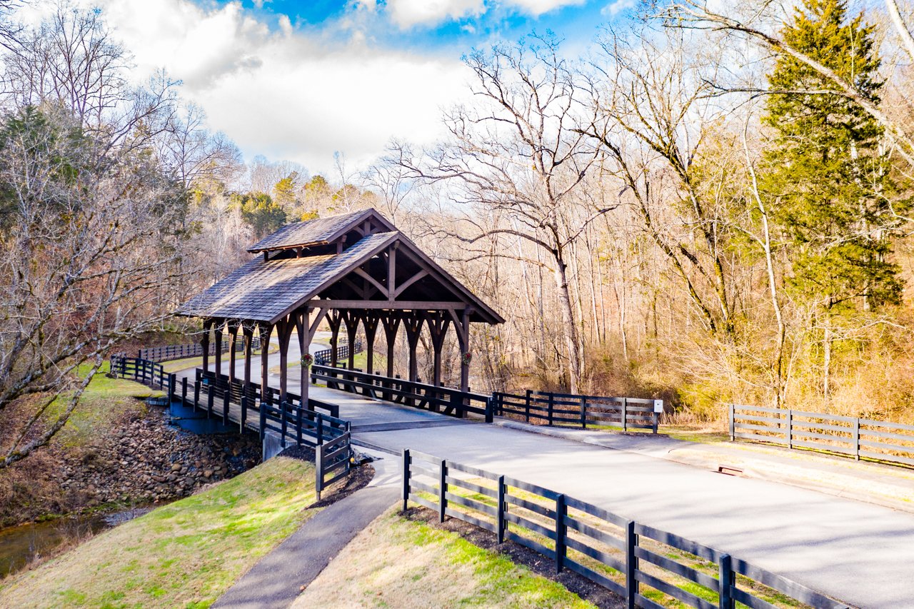 A covered wooden bridge in a rural area during early spring, with leafless trees on either side and a narrow stream running beneath it.
