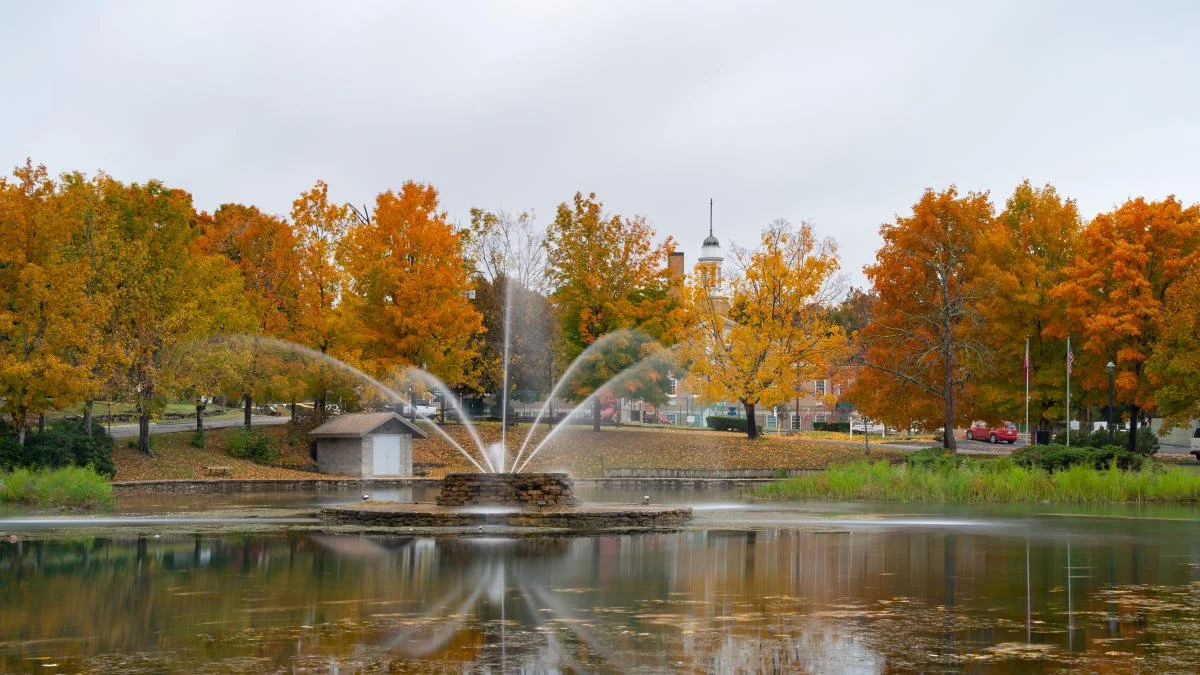 Autumn scene in a park with a pond, a fountain, trees with orange and yellow leaves, and a church in the background.