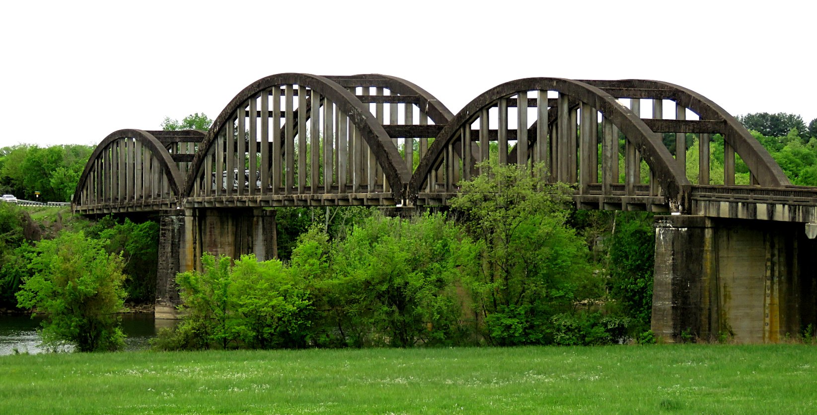 A wooden arch bridge over a river, surrounded by green trees and grass under an overcast sky.