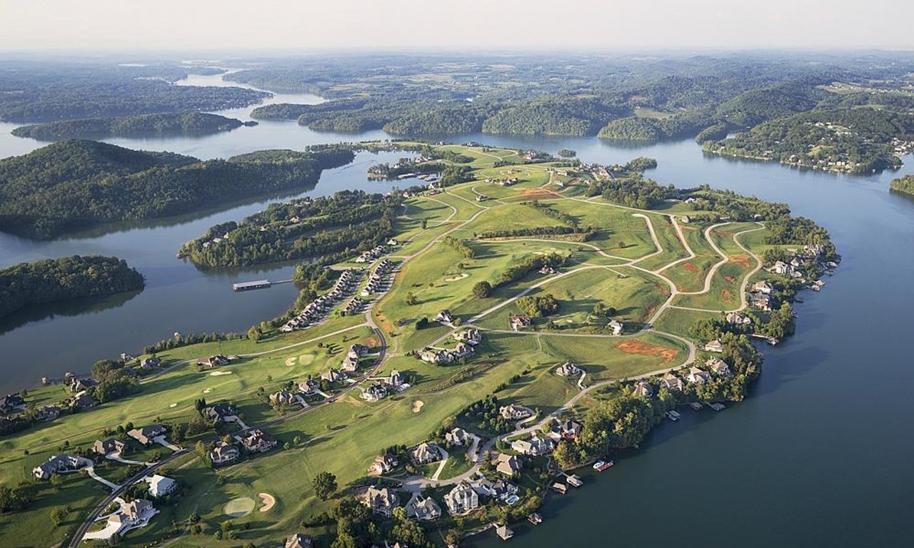 Aerial view of a golf course surrounded by residential homes and water bodies with islands and lush green terrain in the background.