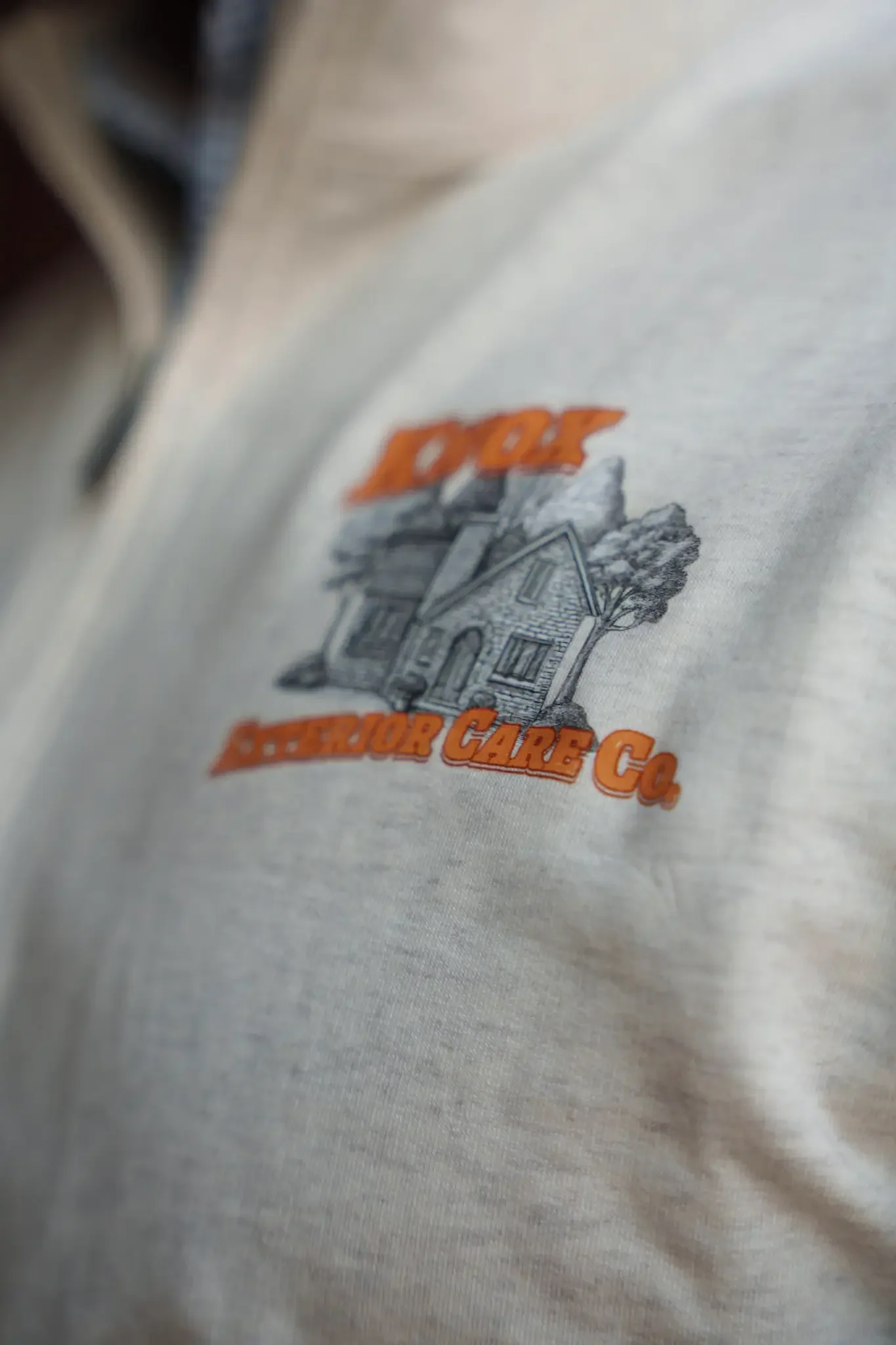 Close-up of a t-shirt with a design featuring a house, trees, and shelter care Co in orange text.