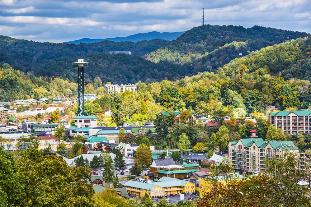 Scenic view of a town in the mountains with colorful buildings, surrounded by green trees and hills, under a partly cloudy sky.